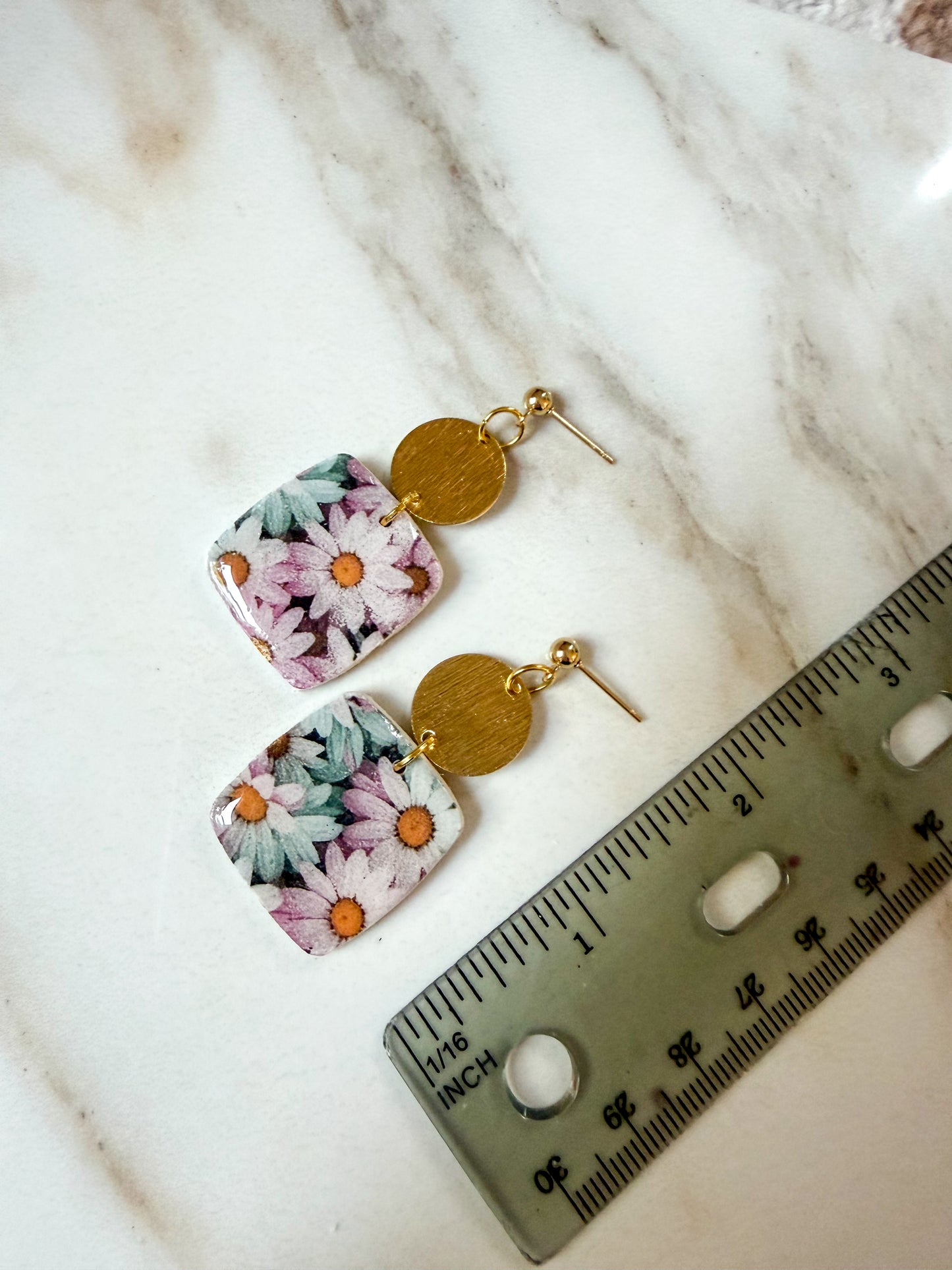 A pair of dangling earrings with floral-patterned square charms and round gold-tone discs, displayed on a white marble surface next to a metal ruler for scale. 2 inches long. 