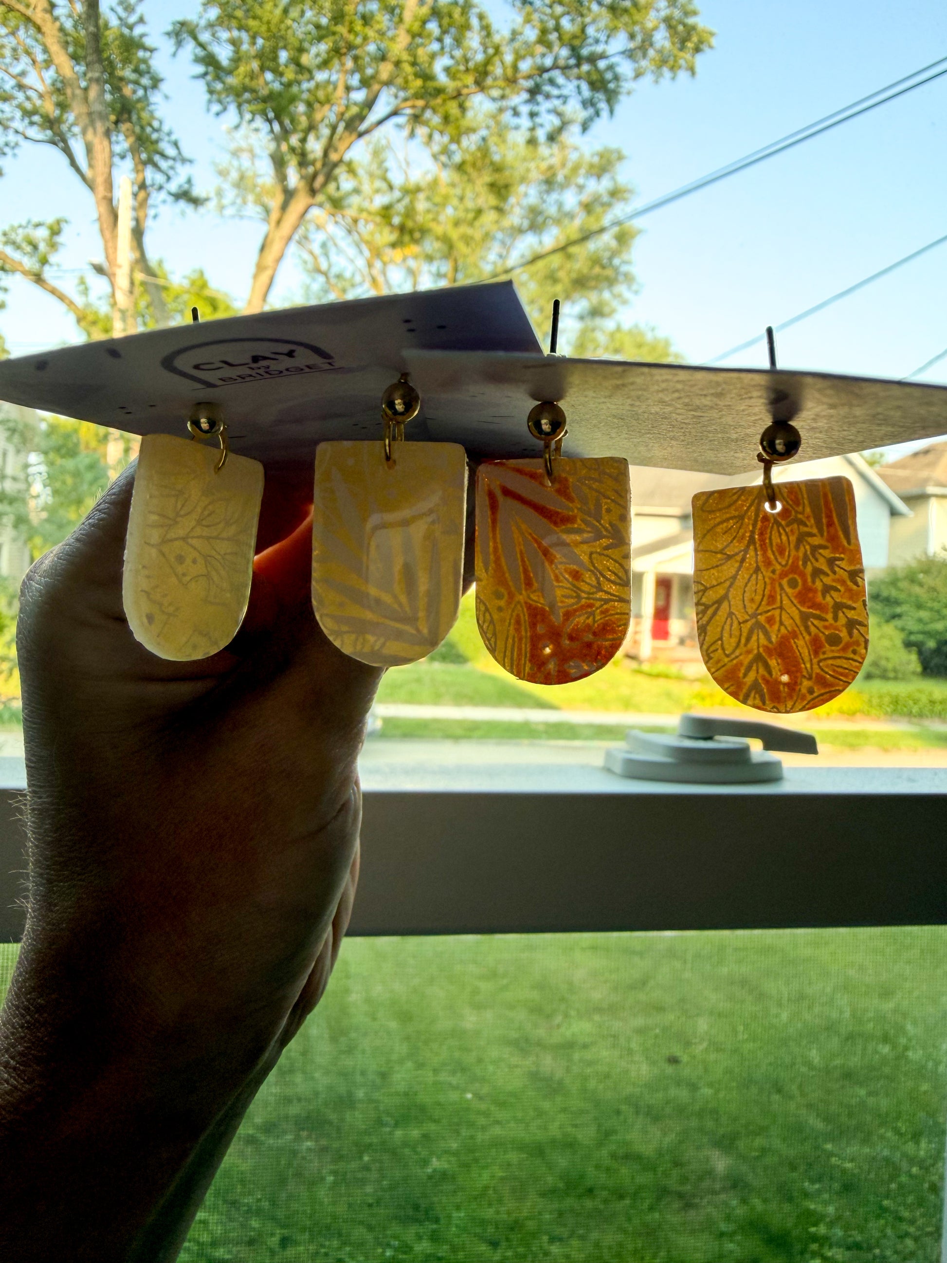 Four drop earrings with gold-toned metal tops and marbled amber, orange, and cream resin pendants, held by a hand in front of a window with a backyard view. 