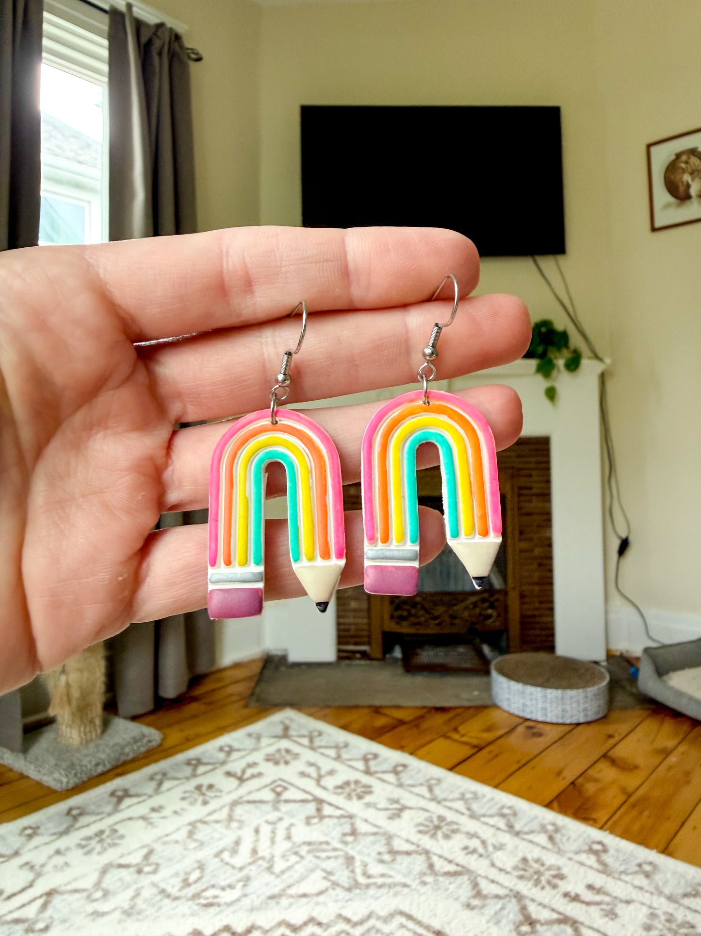 A hand holds a pair of rainbow pencil-shaped drop earrings with silver fishhook wires in a cozy, lived-in room setting.