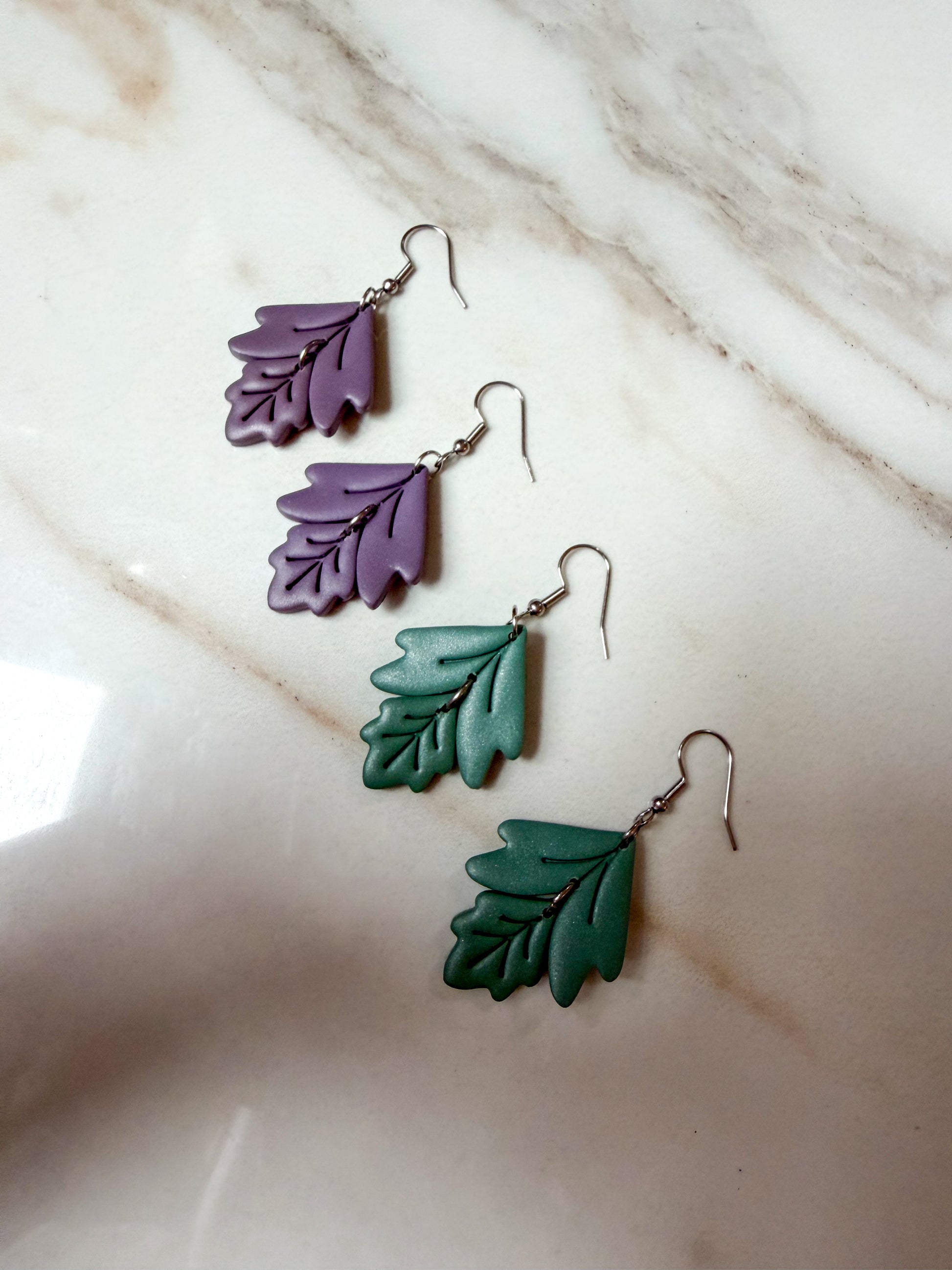 Four leaf-shaped drop earrings in purple and green with silver fishhook wires, arranged on a light beige marble surface.