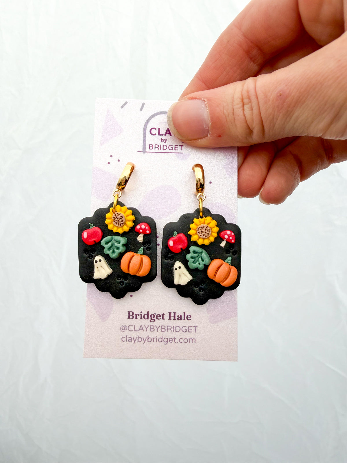 A hand holds a pair of black polymer clay dangle earrings decorated with autumn and Halloween motifs, including sunflowers, pumpkins, ghosts, and mushrooms, on a branded display card.