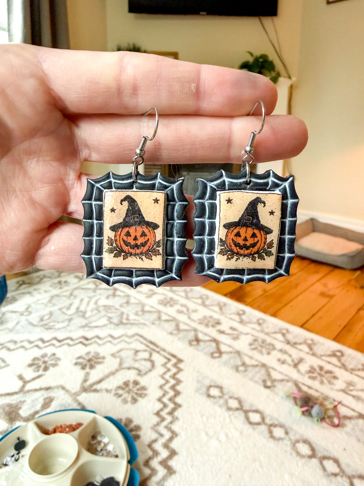 A hand holds a pair of Halloween-themed drop earrings with spiderweb borders and jack-o'-lantern designs, shown in a home setting.