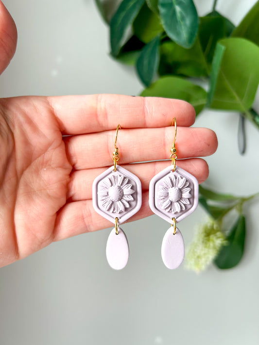 Purple monochromatic hexagon shaped earrings with a daisy in the center. Held in a hand. 