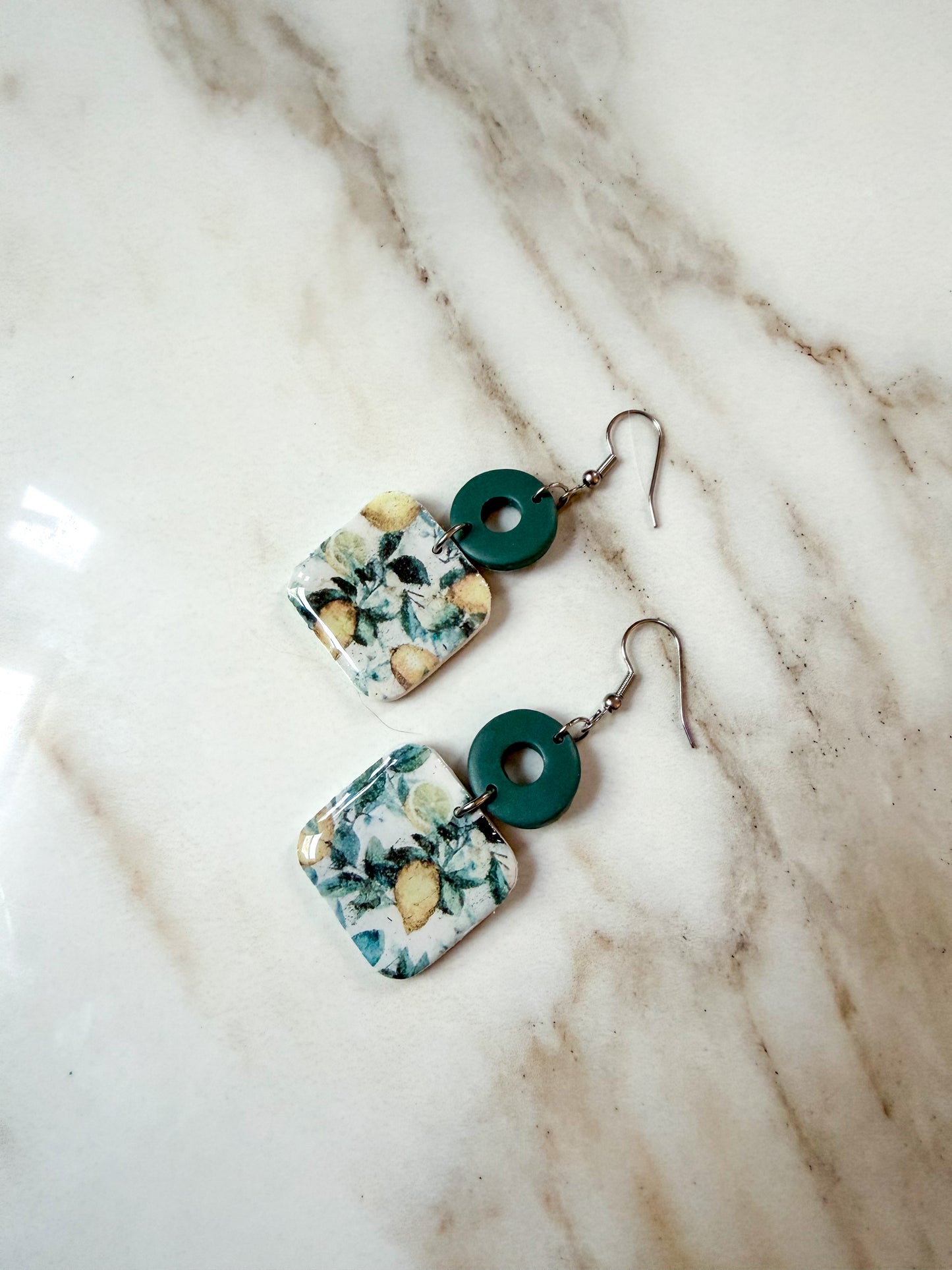 A pair of handmade dangle earrings featuring square floral resin pendants in teal, white, and yellow, suspended from dark green circular rings on silver fishhook wires, displayed on a white marble surface.