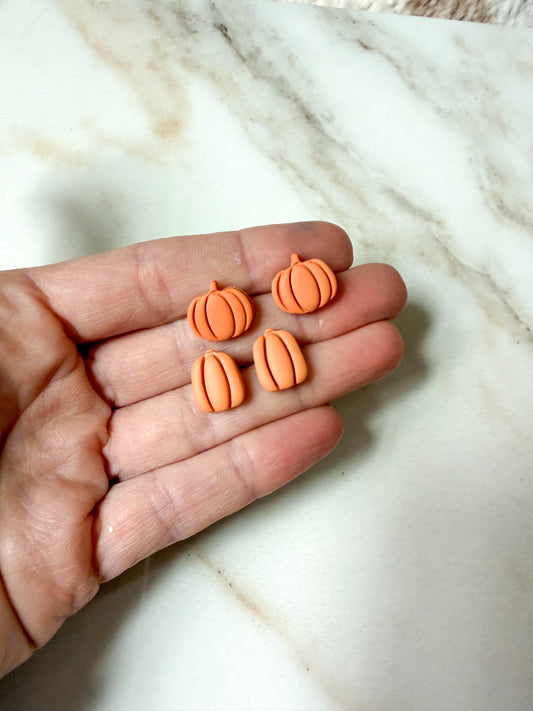 Four small orange pumpkin-shaped stud earrings held in an open hand over a white marble surface.