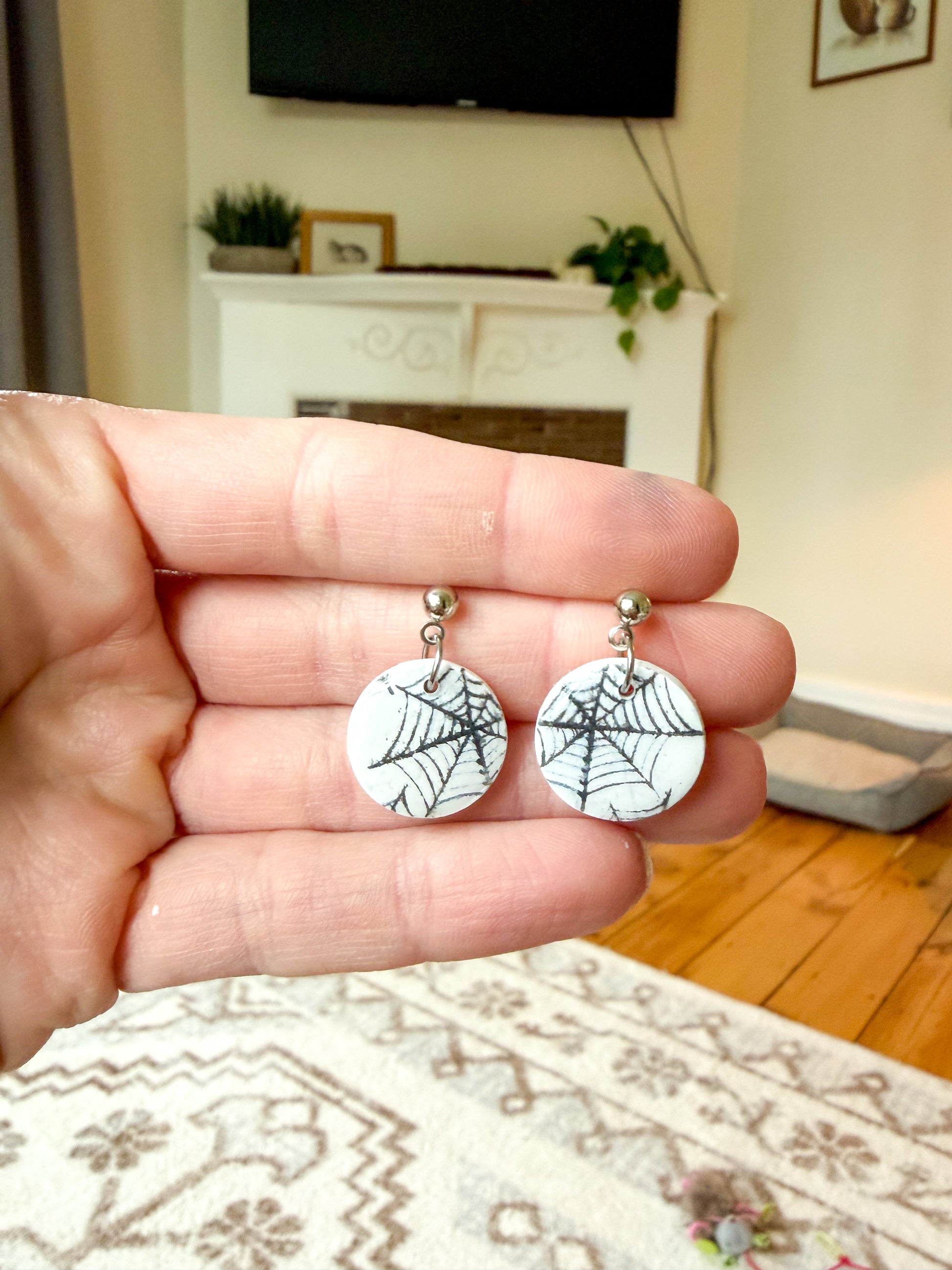 A hand holding a pair of round white earrings with black spiderweb designs, shown in a home interior setting with a fireplace and wooden floor in the background.
