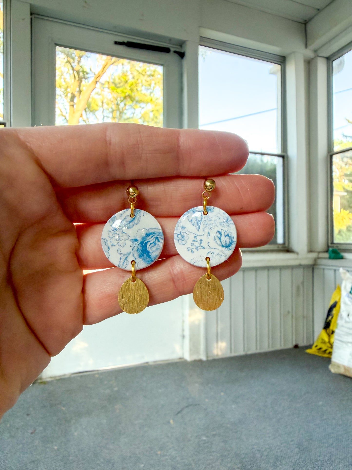Hand holding a pair of earrings with a window and outdoor view in the background
