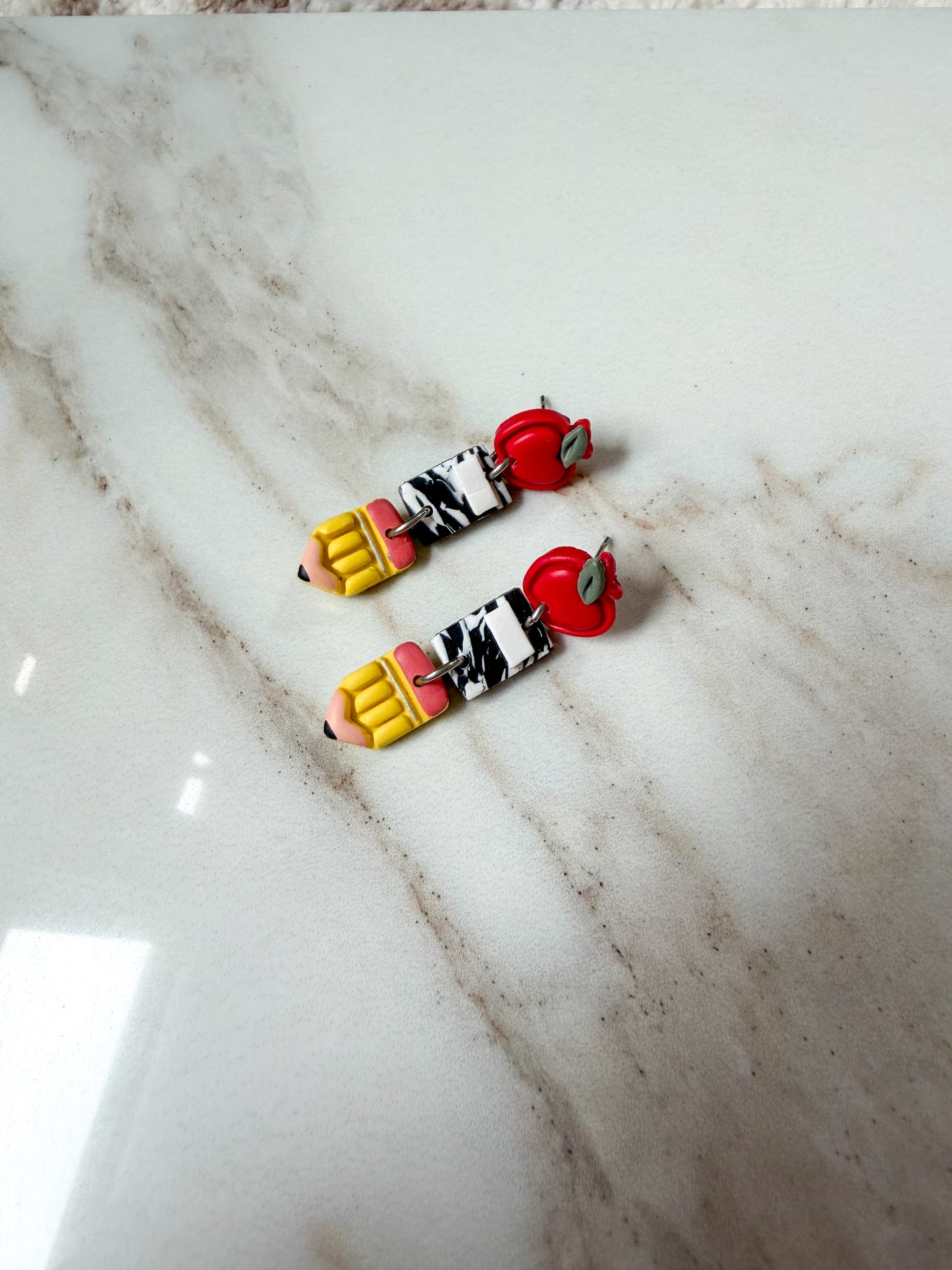 A pair of handmade polymer clay earrings on a white marble surface. Apples on top, with composition notebooks in the middle, and pencils at the bottom. 