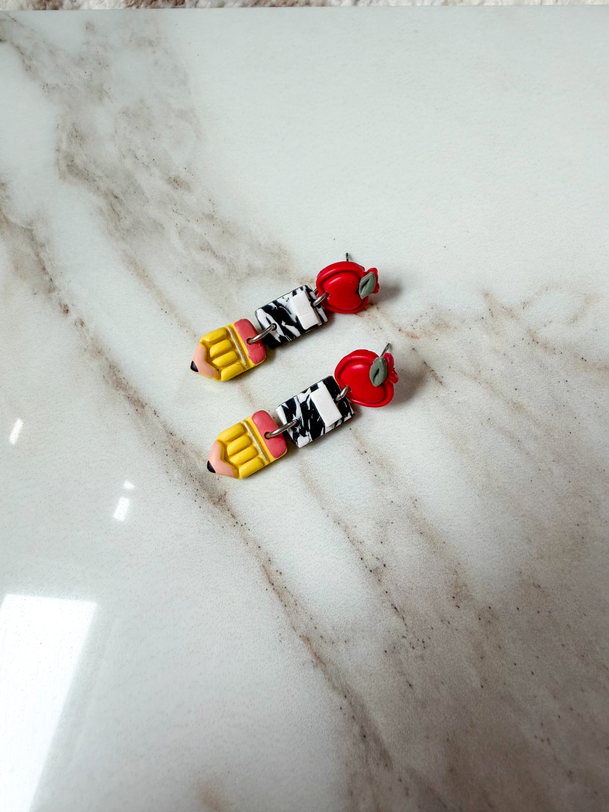 A pair of handmade polymer clay earrings on a white marble surface. Apples on top, with composition notebooks in the middle, and pencils at the bottom. 