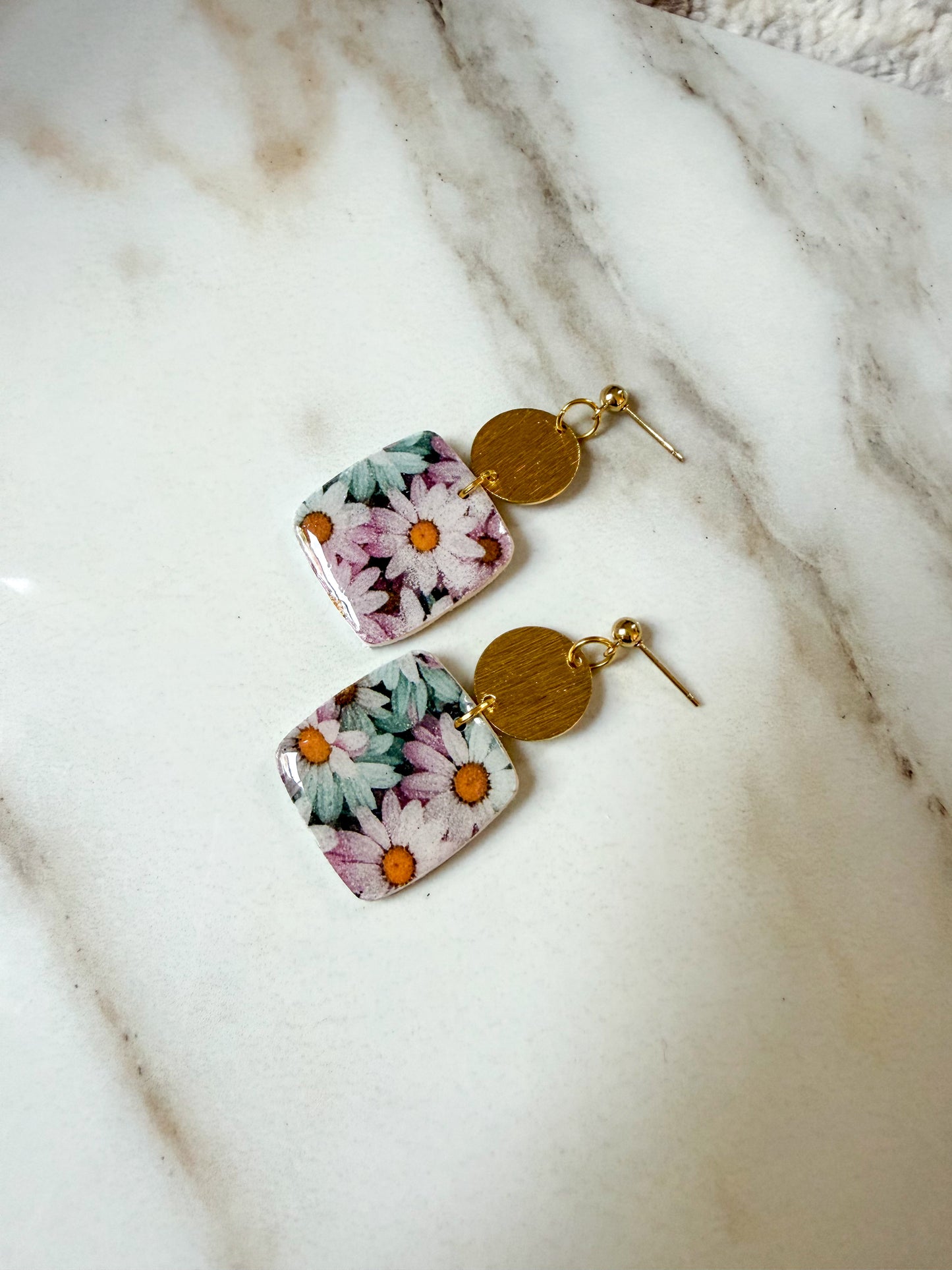 A pair of square daisy-patterned earrings with gold disc charms, displayed on a white marble surface.