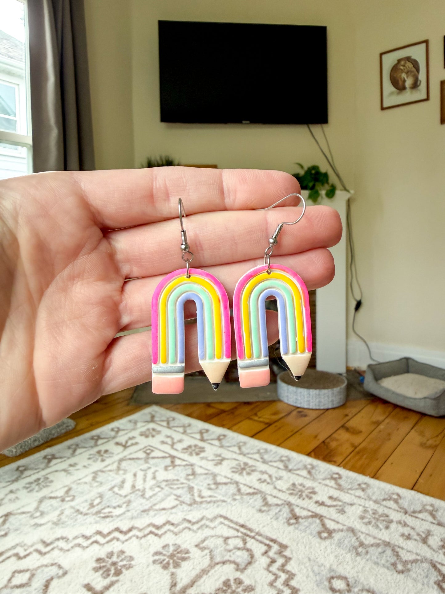 A pair of rainbow-and-pencil drop earrings held in a person's hand, with a real living room background including a TV, rug, and floor.