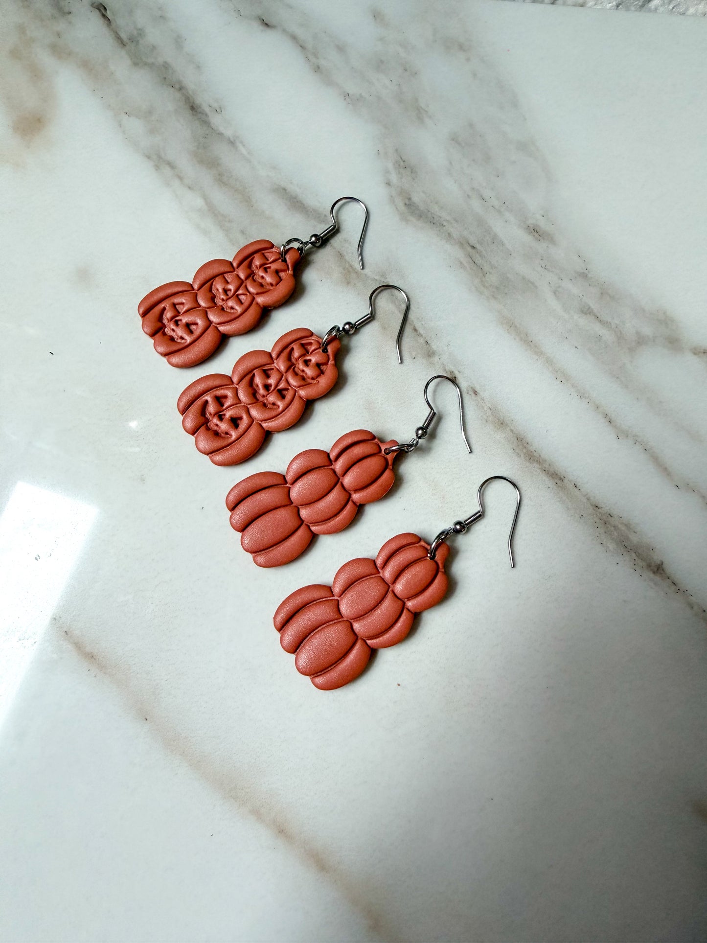 Two pairs of orange pumpkin-shaped dangle earrings with silver fishhook wires, arranged diagonally on a white marble surface.