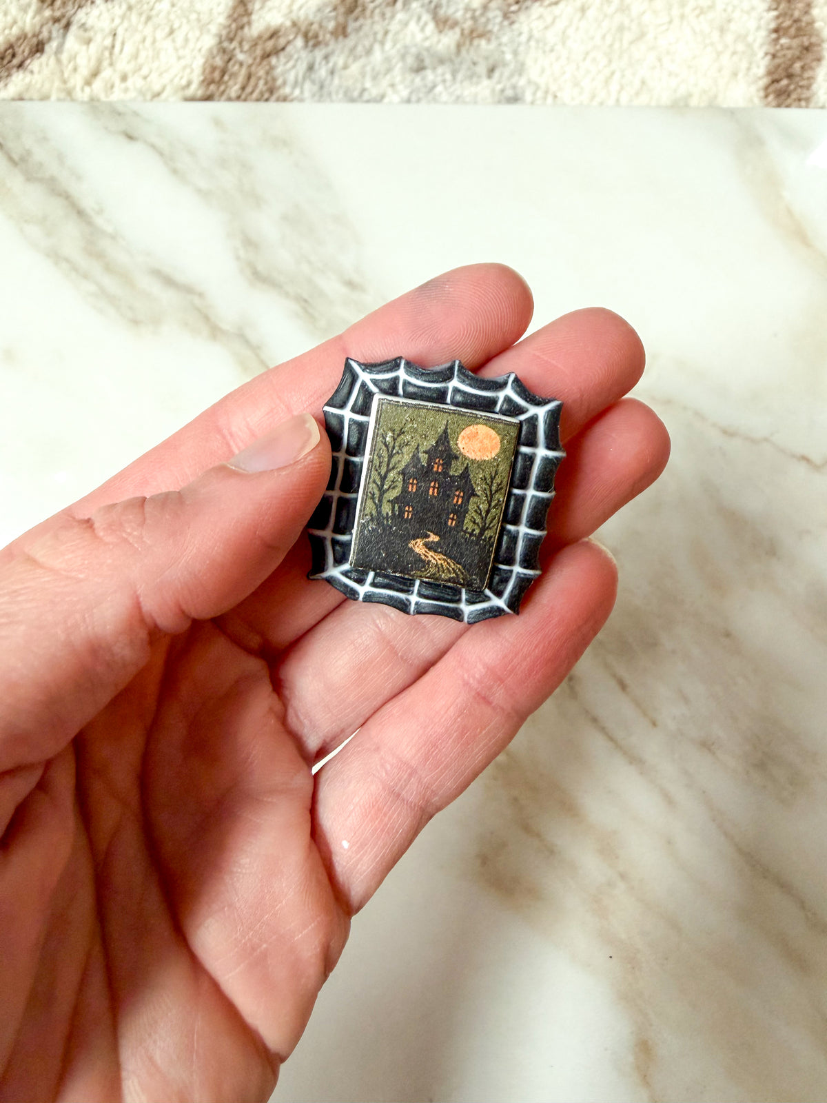 A hand holding a small square Halloween-themed magnet featuring a haunted house, spiderweb border, and full moon on a marble surface.