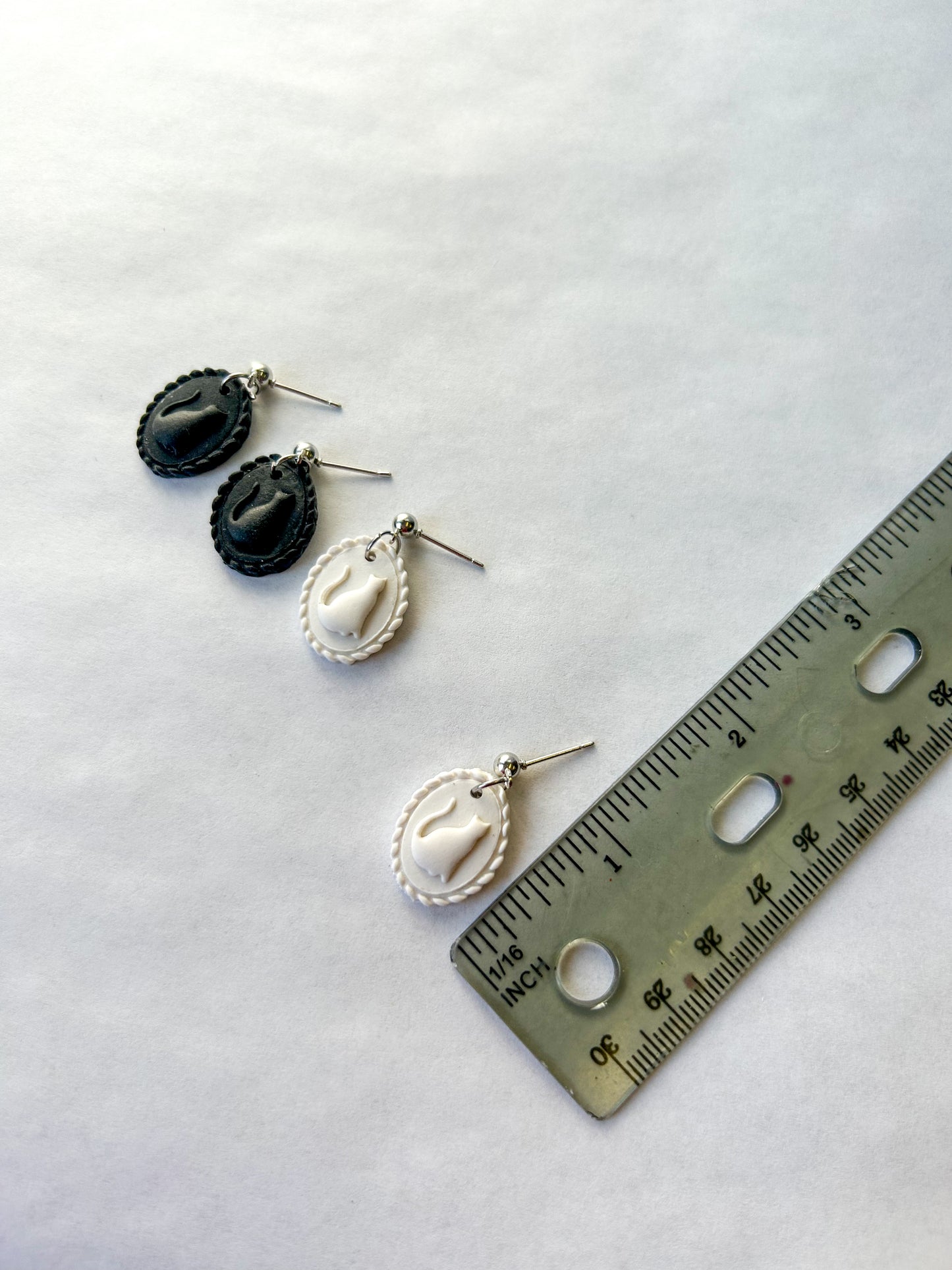 Two pairs of earrings on a white surface with a ruler for scale. The earrings resemble a scalloped frame with a raised cat silhouette in the center.