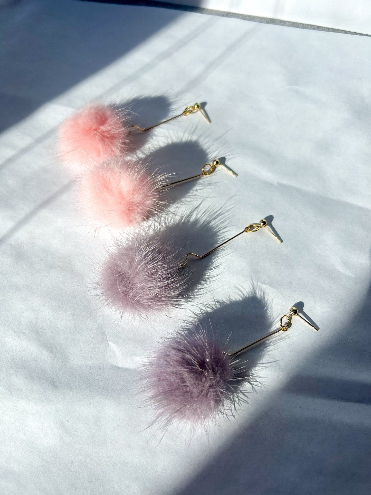 Earrings laying on a flat surface. One pair of pink and one pair of purple. They have a fuzzy ball at the bottom with a gold squiggly metal dangle. 