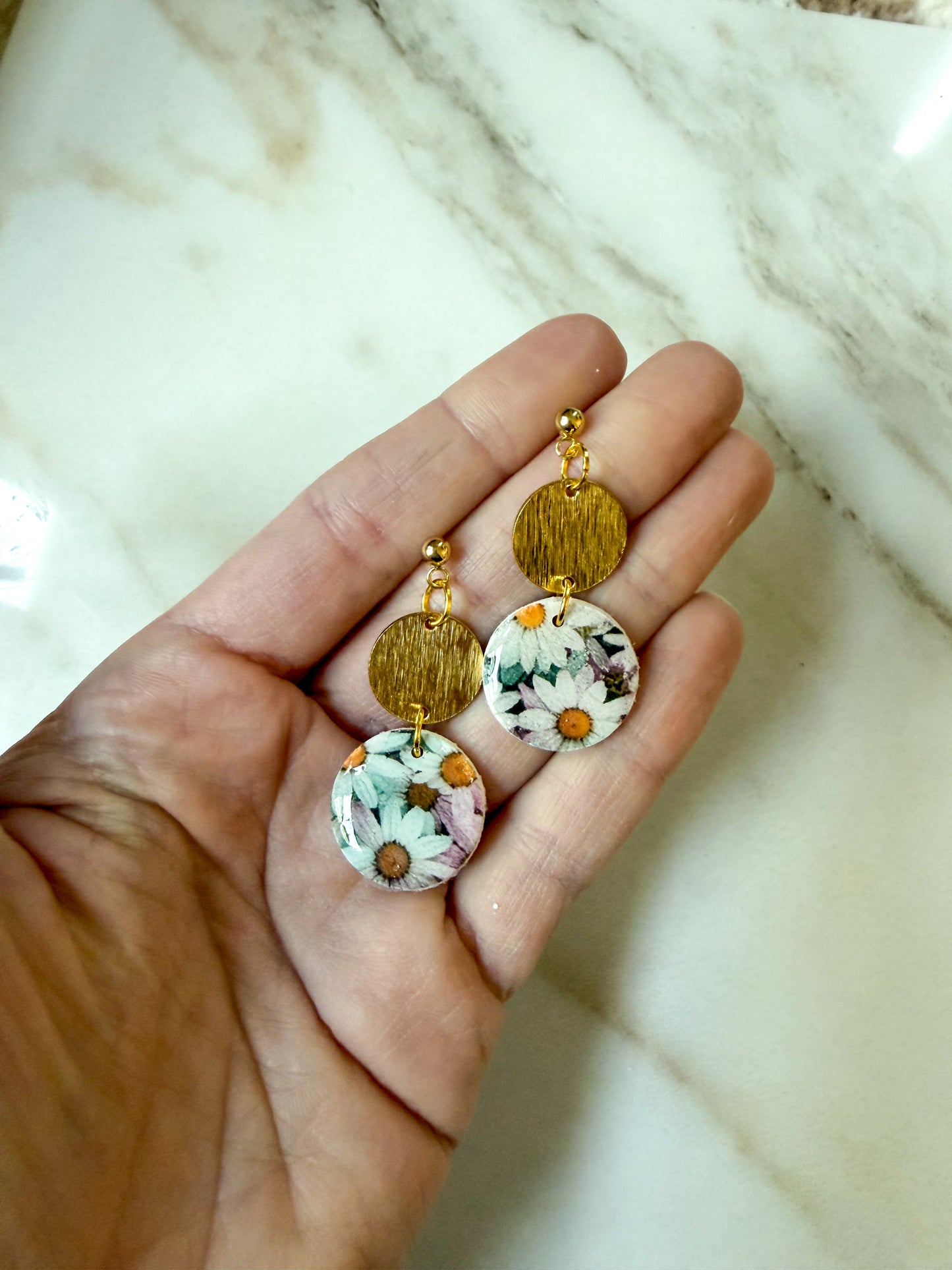 A pair of handmade dangle earrings held in a hand. The earrings feature textured gold-tone circular discs at the top and floral-patterned round drops at the bottom with white, pink, and yellow flowers on a light background.