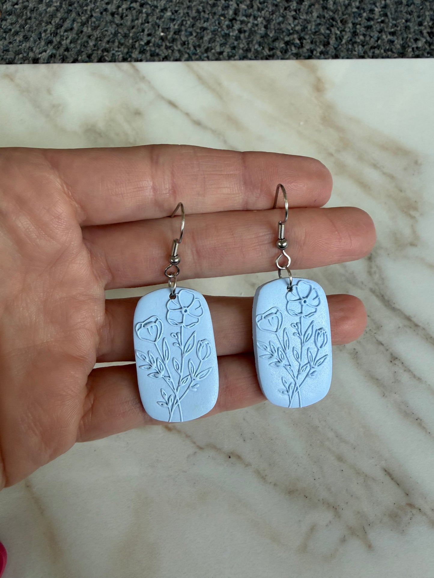 Hand holding two blue earrings with floral carvings on a marble surface.