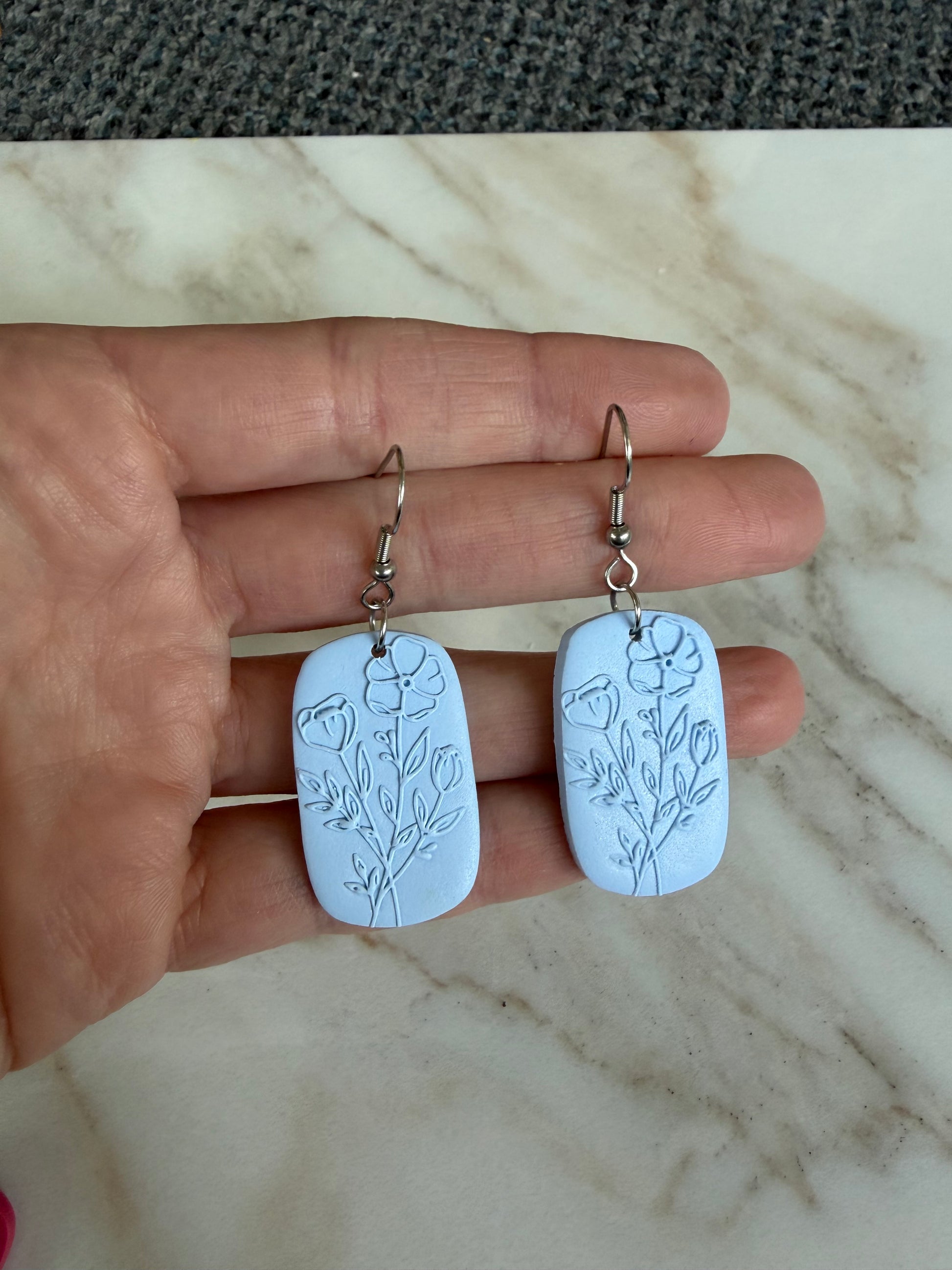 Hand holding two blue earrings with floral carvings on a marble surface.