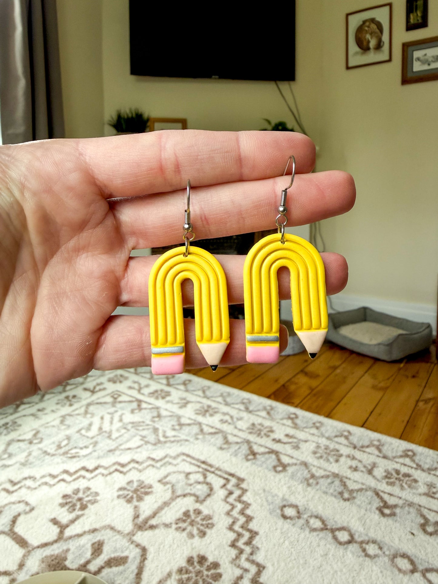 A hand holds a pair of yellow pencil-shaped earrings with silver hooks in a home setting with a patterned rug and wooden floor in the background.