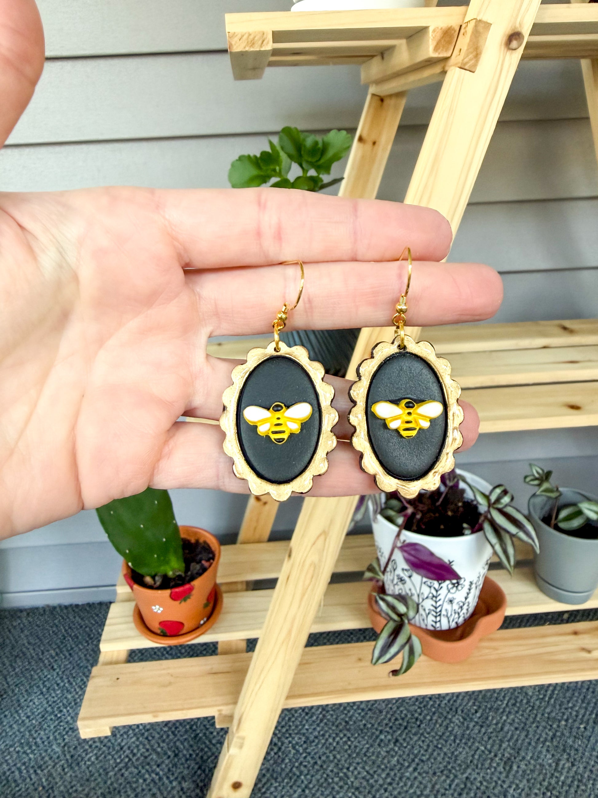 Hand holding a pair of earrings with bee design against a wooden stand and plant background