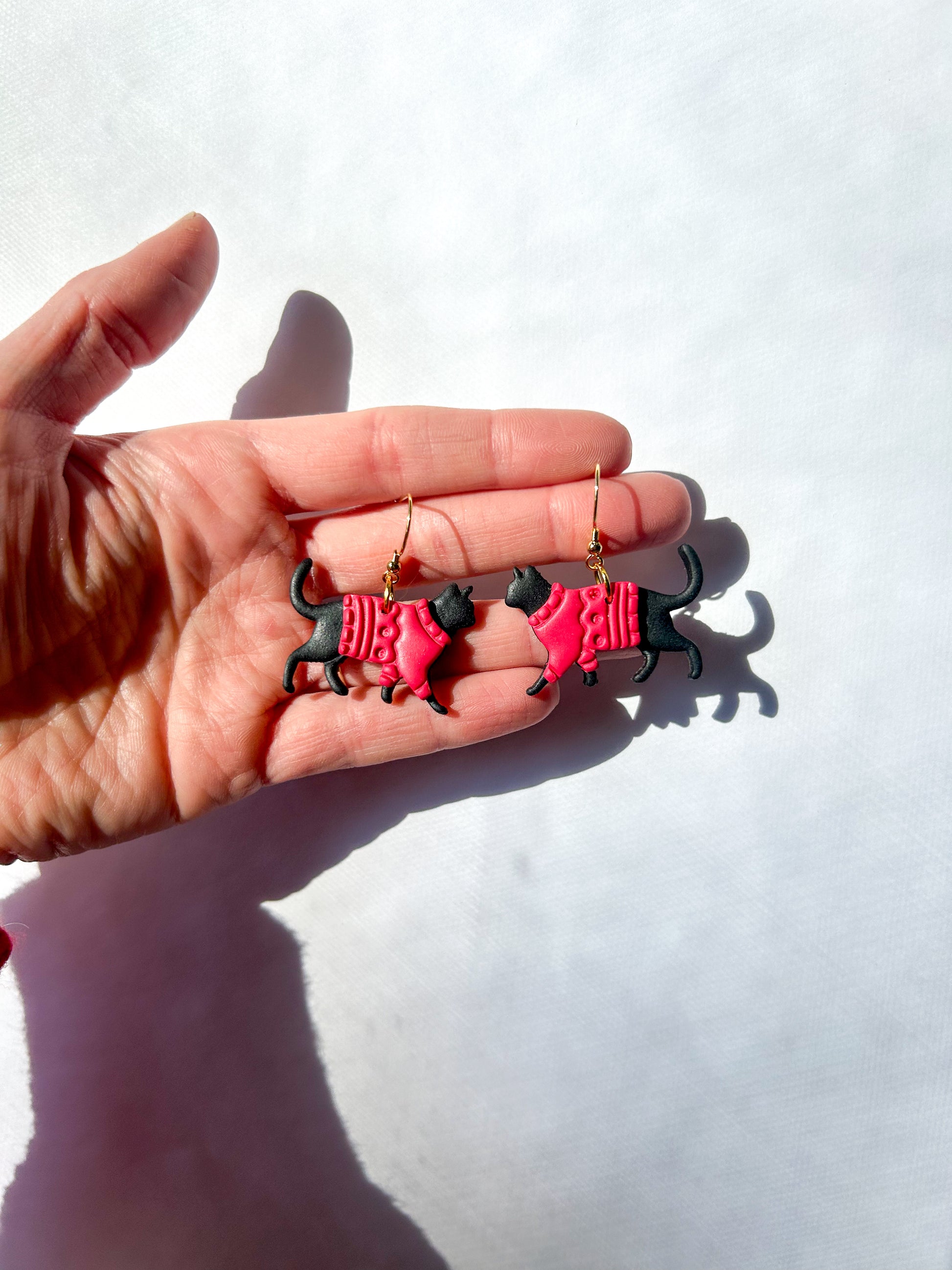 Hand holding two black cat-shaped earrings against a white background. The cats have red sweaters. 