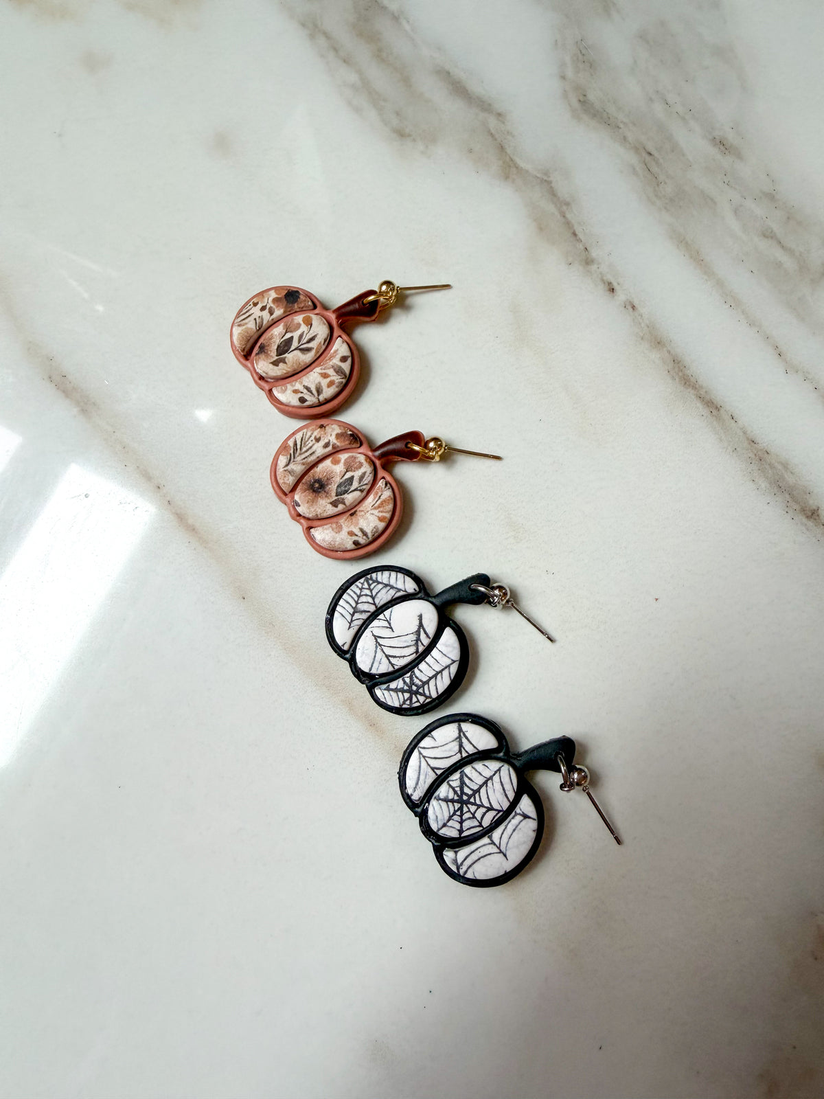 Two pairs of pumpkin shaped earrings with decorative designs laying on a marble surface. 