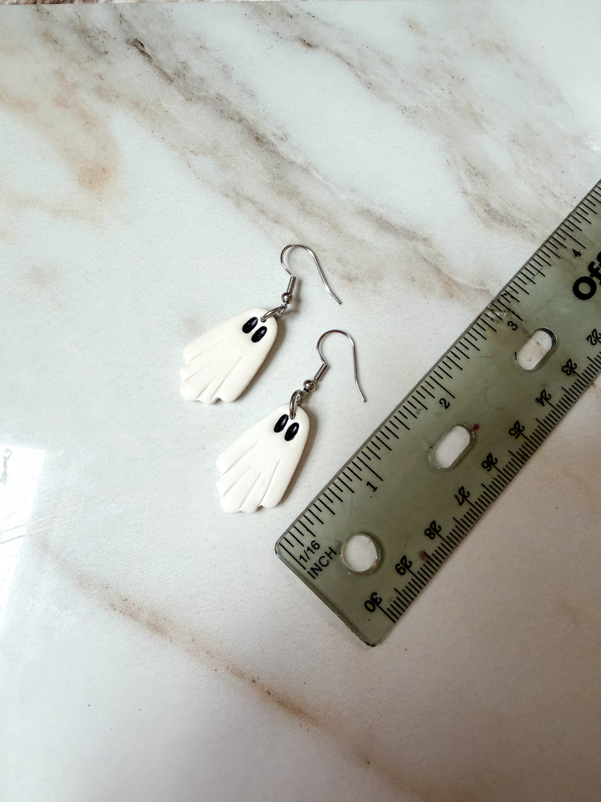 A pair of white ghost-shaped dangle earrings with black eyes and silver fishhook wires, displayed on a white marble surface next to a ruler for scale. About 2 inches long. 