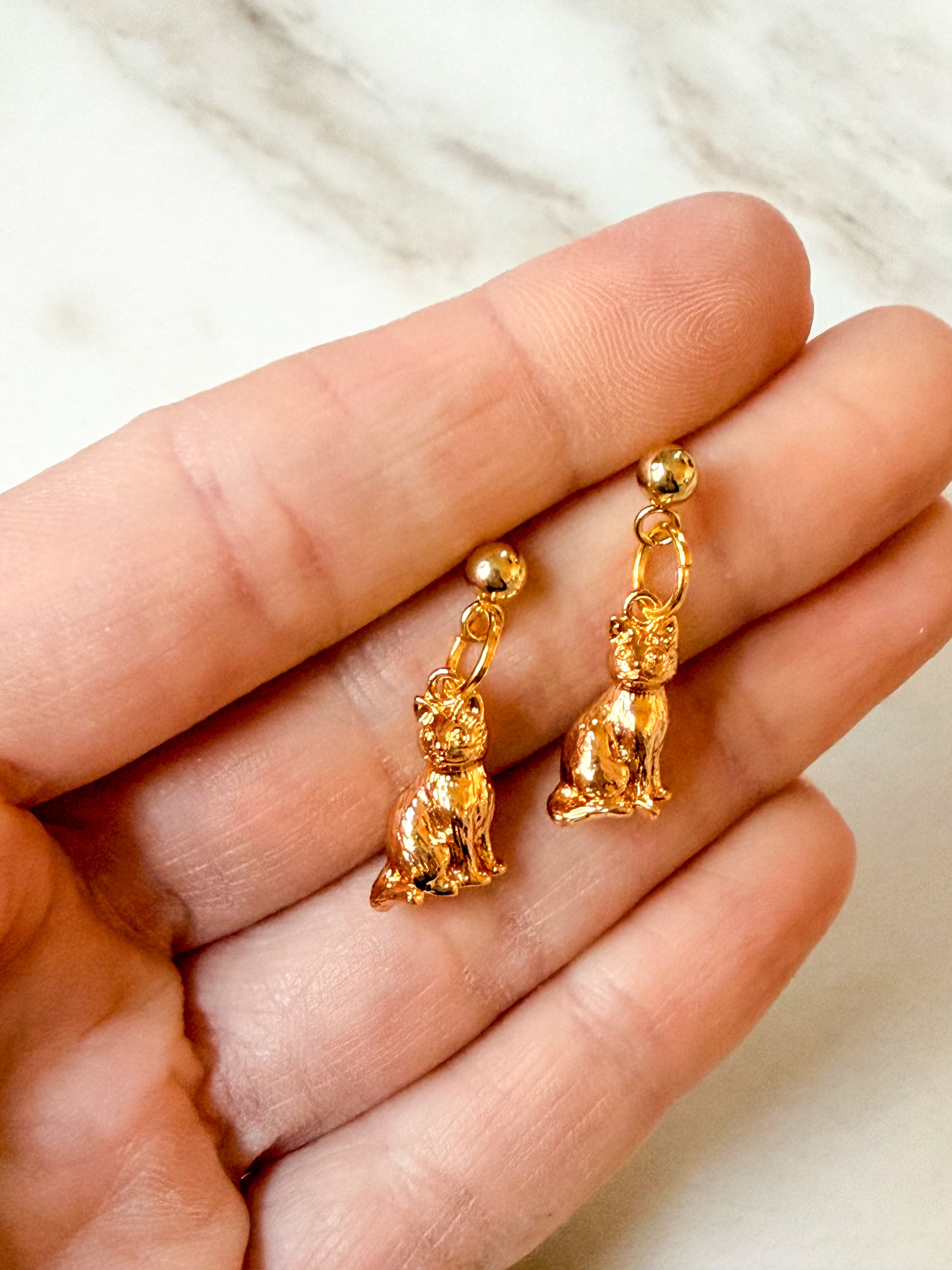 A pair of small gold-tone dangling earrings shaped like seated cats, held between a person's fingers against a light marble-like background.