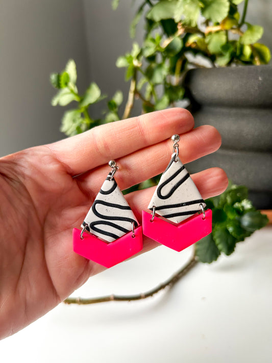 Kite shaped dangles with a chevron bottom piece that is attached separately. The top is white with a black swirl design over it. The chevron bottom is hot pink. 