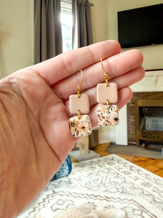 A hand holds a pair of dangling earrings in front of a living room background. The earrings have gold-tone hooks and square charms, with one charm being a solid pale peach-beige and the other featuring a floral print in beige, brown, and white.