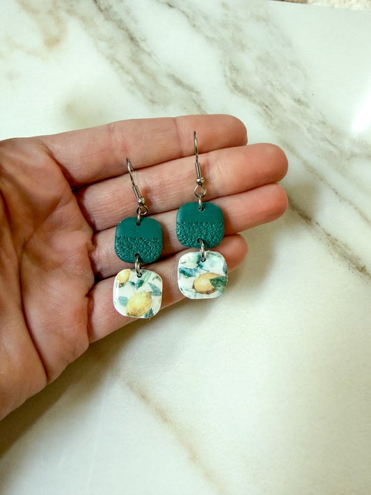 A pair of handmade dangle earrings held in a hand against a light marble background. The earrings feature textured teal green square charms and white square charms with a yellow and green lemon pattern, hanging from silver fishhook wires.