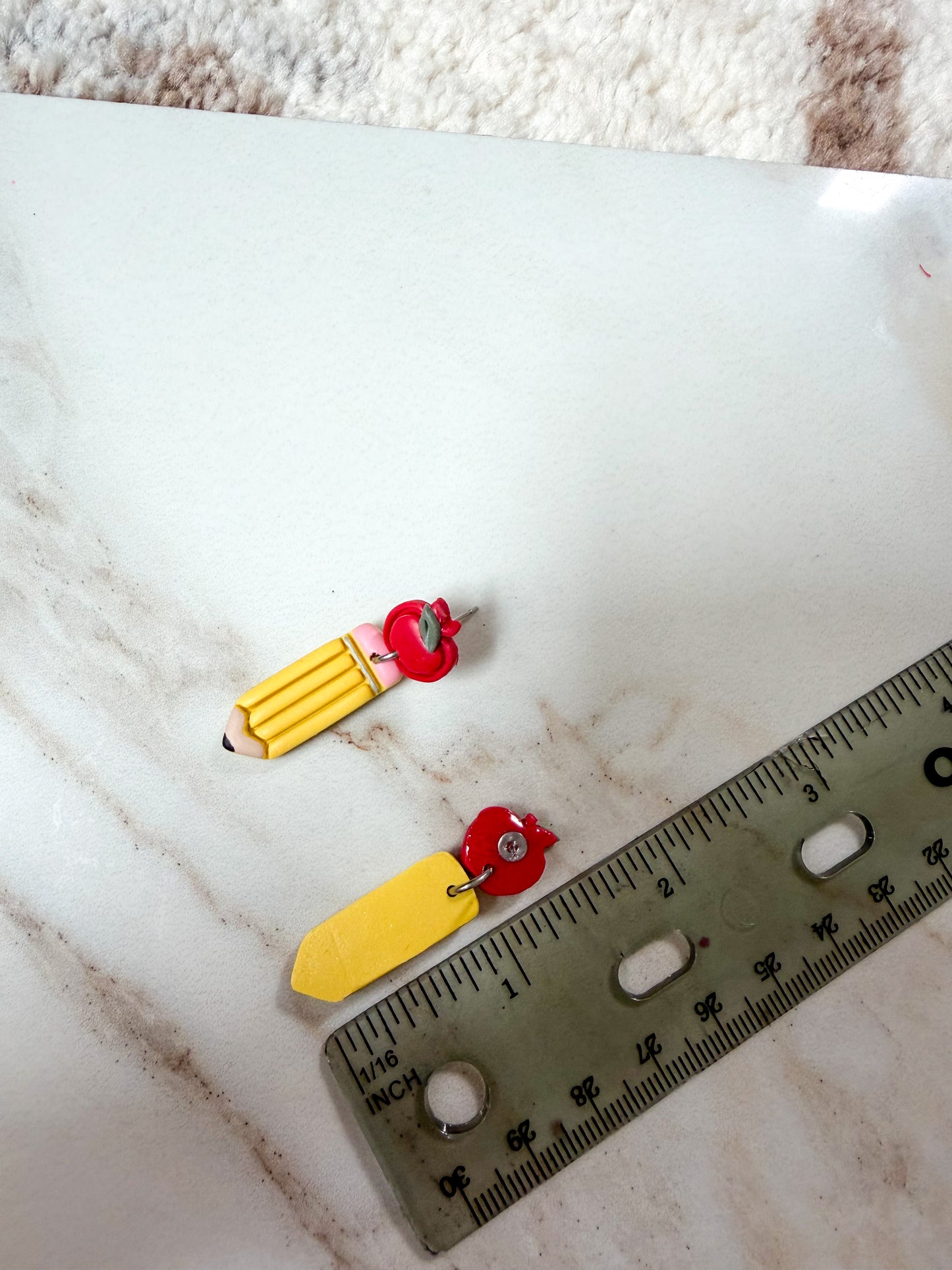 Two small novelty earrings shaped like pencils featuring red apple charms, displayed on a white marble surface next to a ruler for scale. About 1 1/2 inches long. 