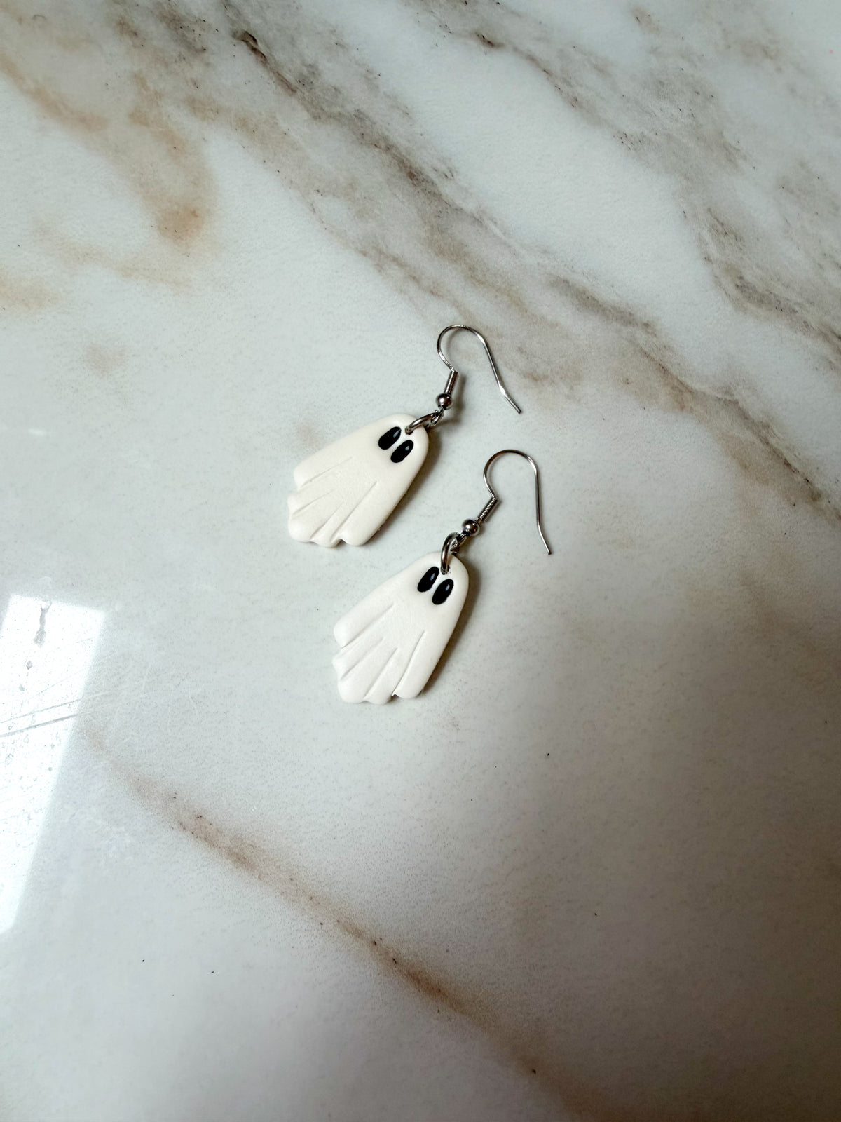 A pair of white ghost-shaped dangle earrings with black eyes and silver fishhook wires, resting on a white and gray marble surface.