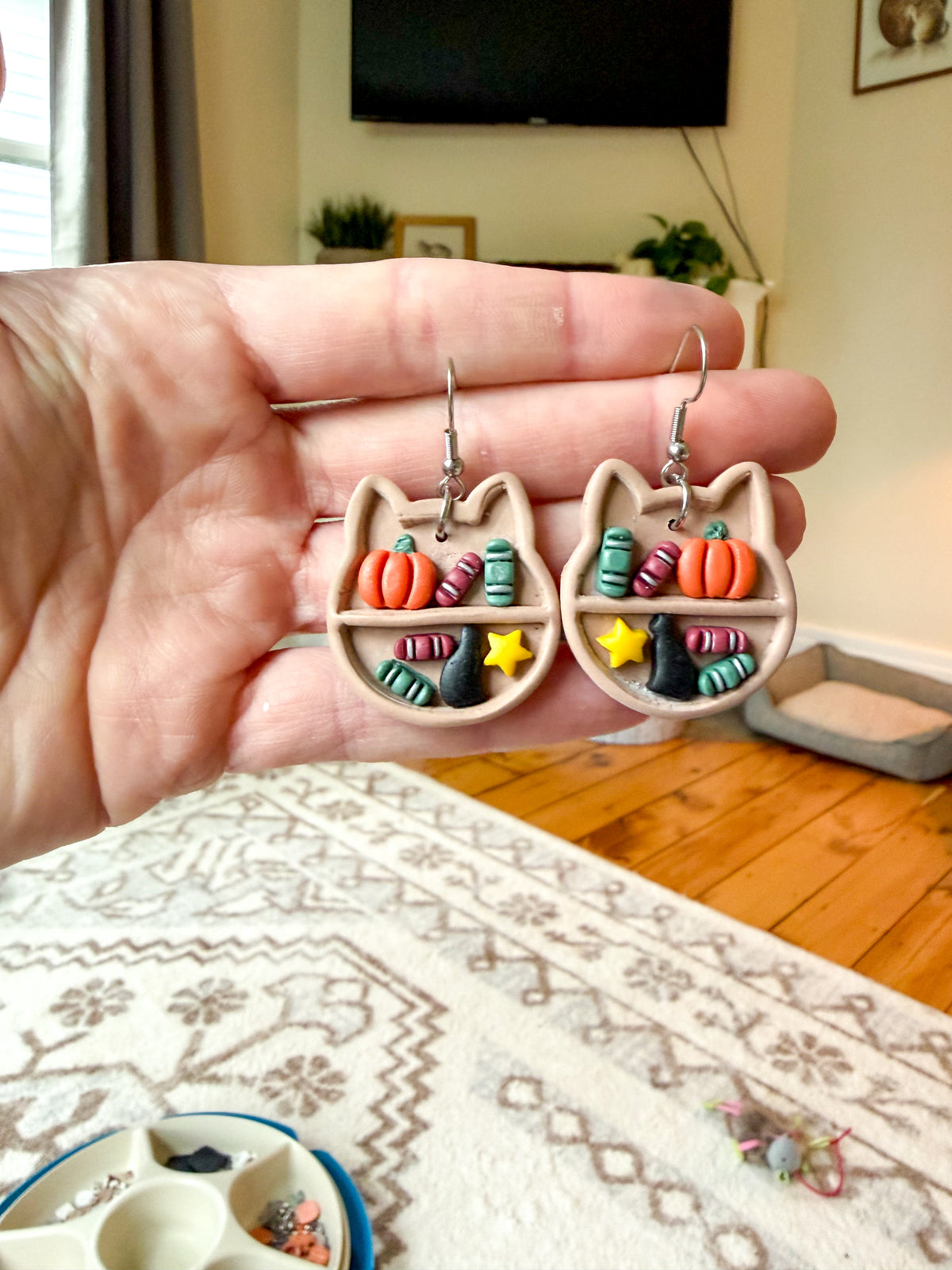 Hand holding earrings that resemble a bookshelf in a cat head shape. The shelves have a black cat and an assortment of books on them. Held against a living room background. 