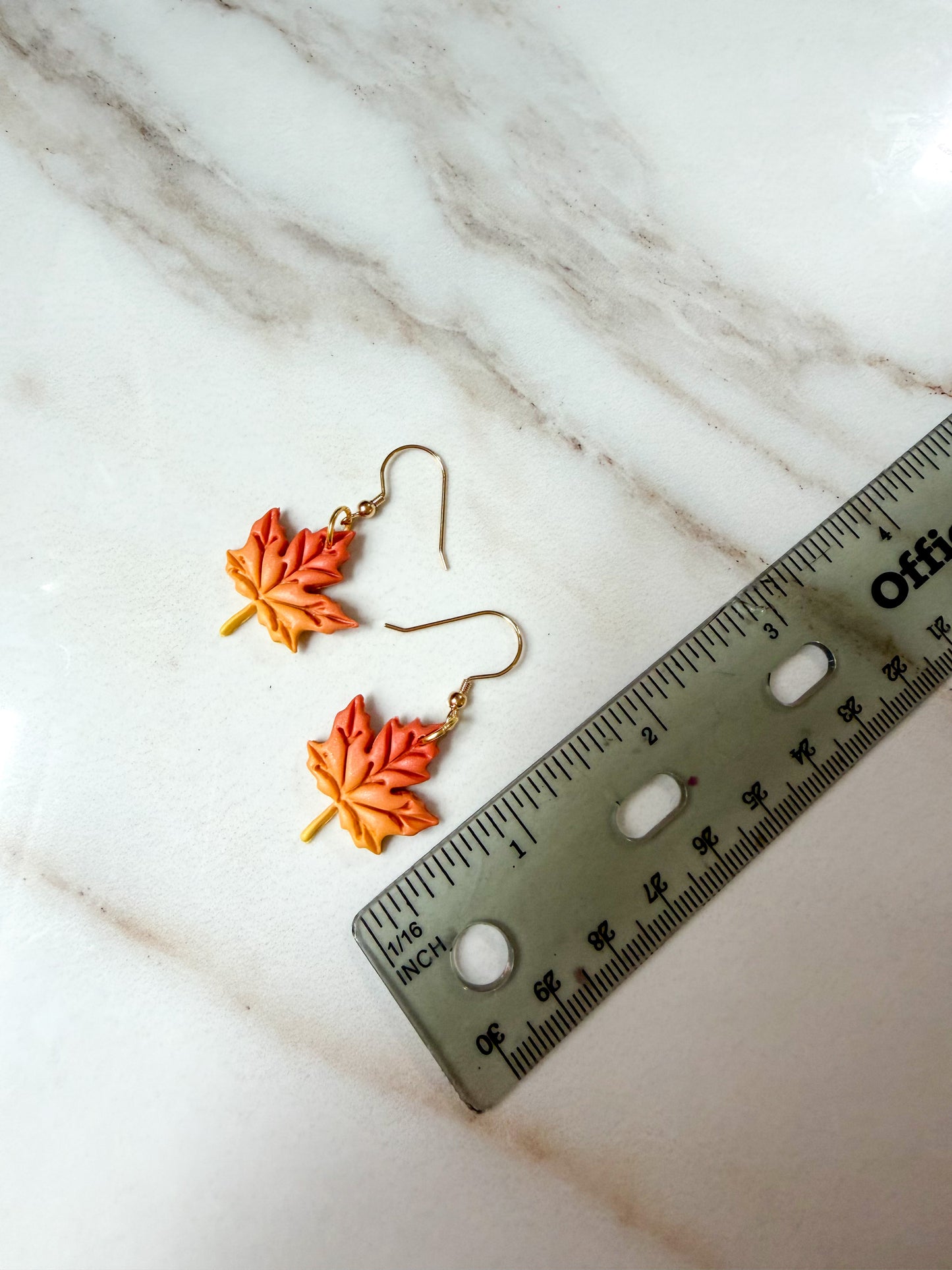 A pair of autumn maple leaf earrings in orange and yellow with gold-tone hooks, displayed on a white marble surface next to a ruler for scale. About 2 inches in length. 