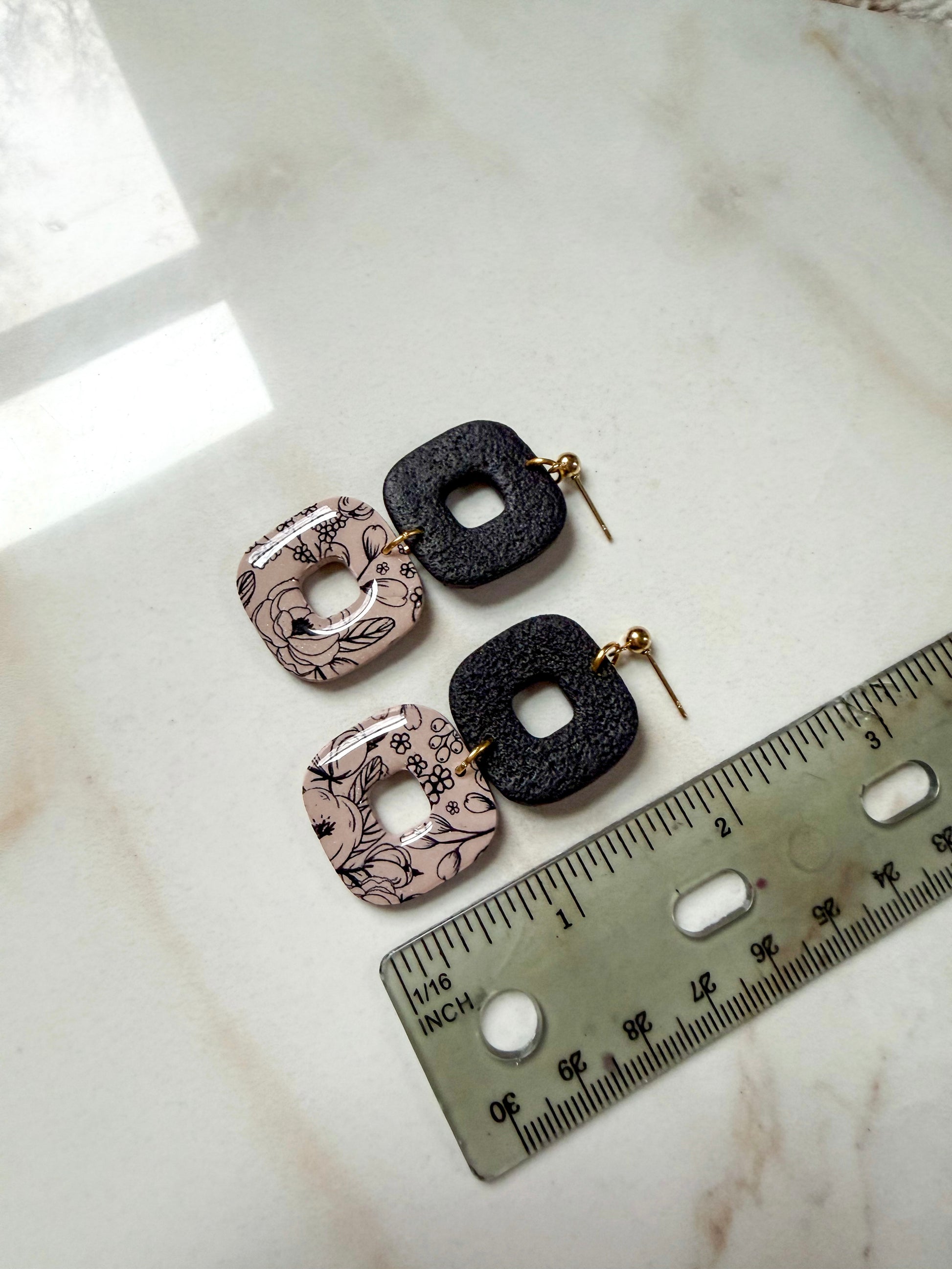 Pair of earrings with floral patterns on a marble surface next to a ruler for scale. About 2 inches long. 