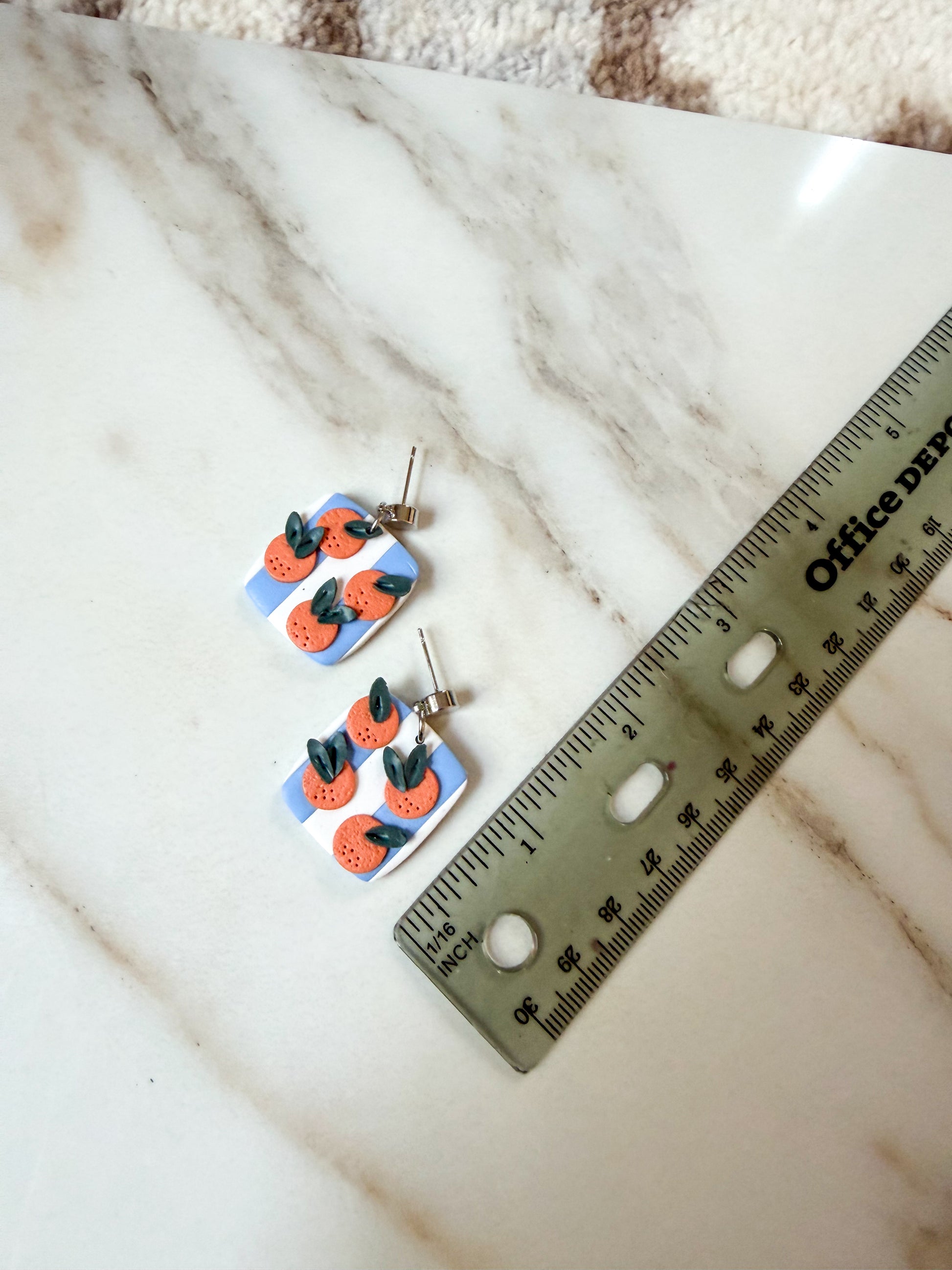 A pair of square earrings with a blue and white striped background, orange fruit illustrations, and green leaves, shown on a white marble surface next to a ruler for scale. About 1 inch long. 
