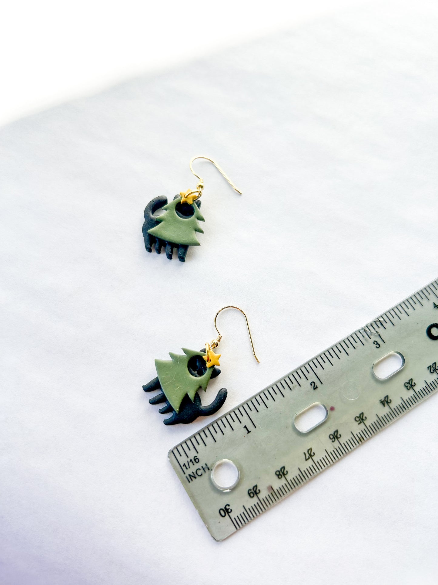Earrings on a white background with a ruler for scale. They are black cats wearing green trees costumes