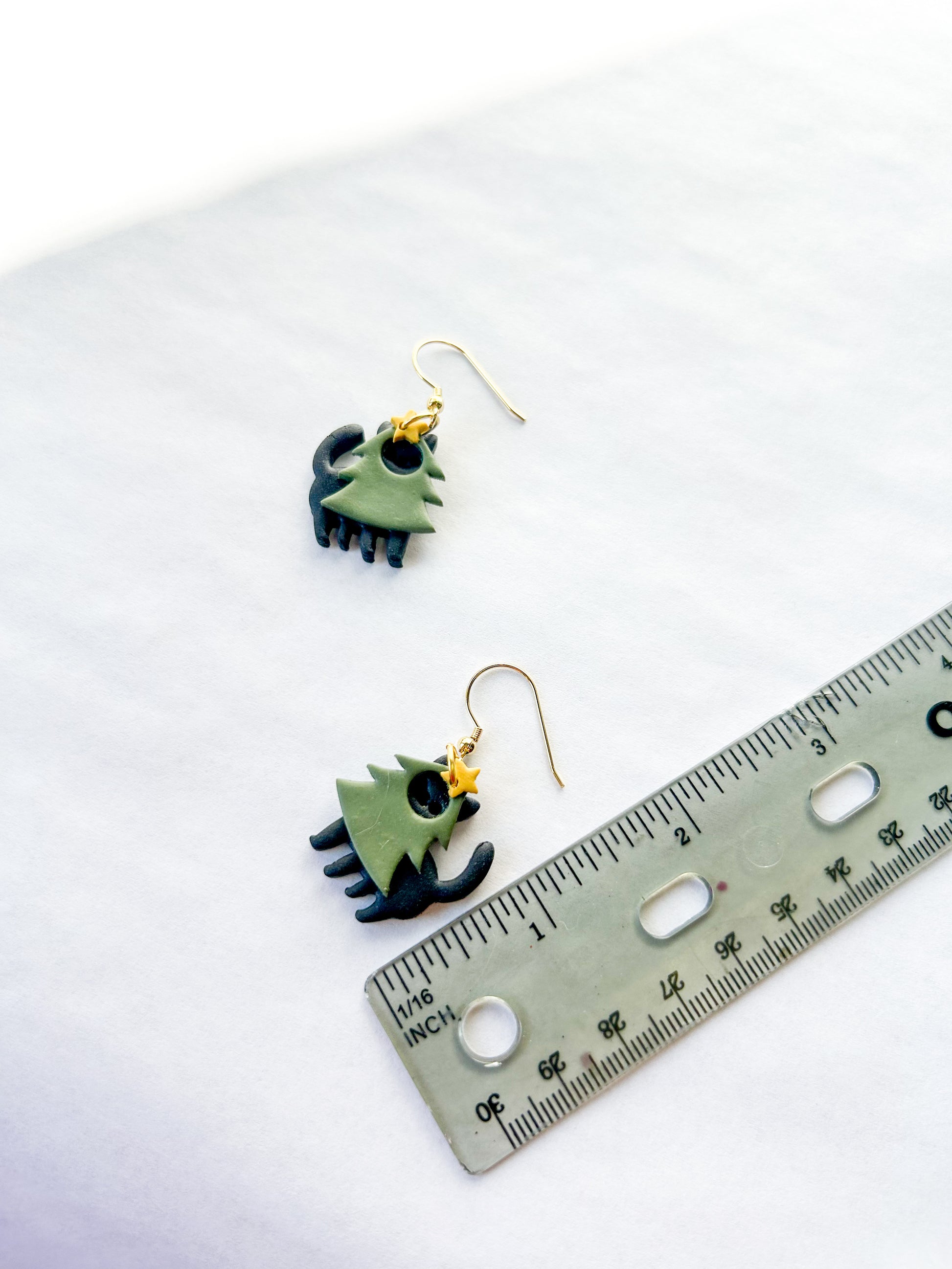 Earrings on a white background with a ruler for scale. They are black cats wearing green trees costumes