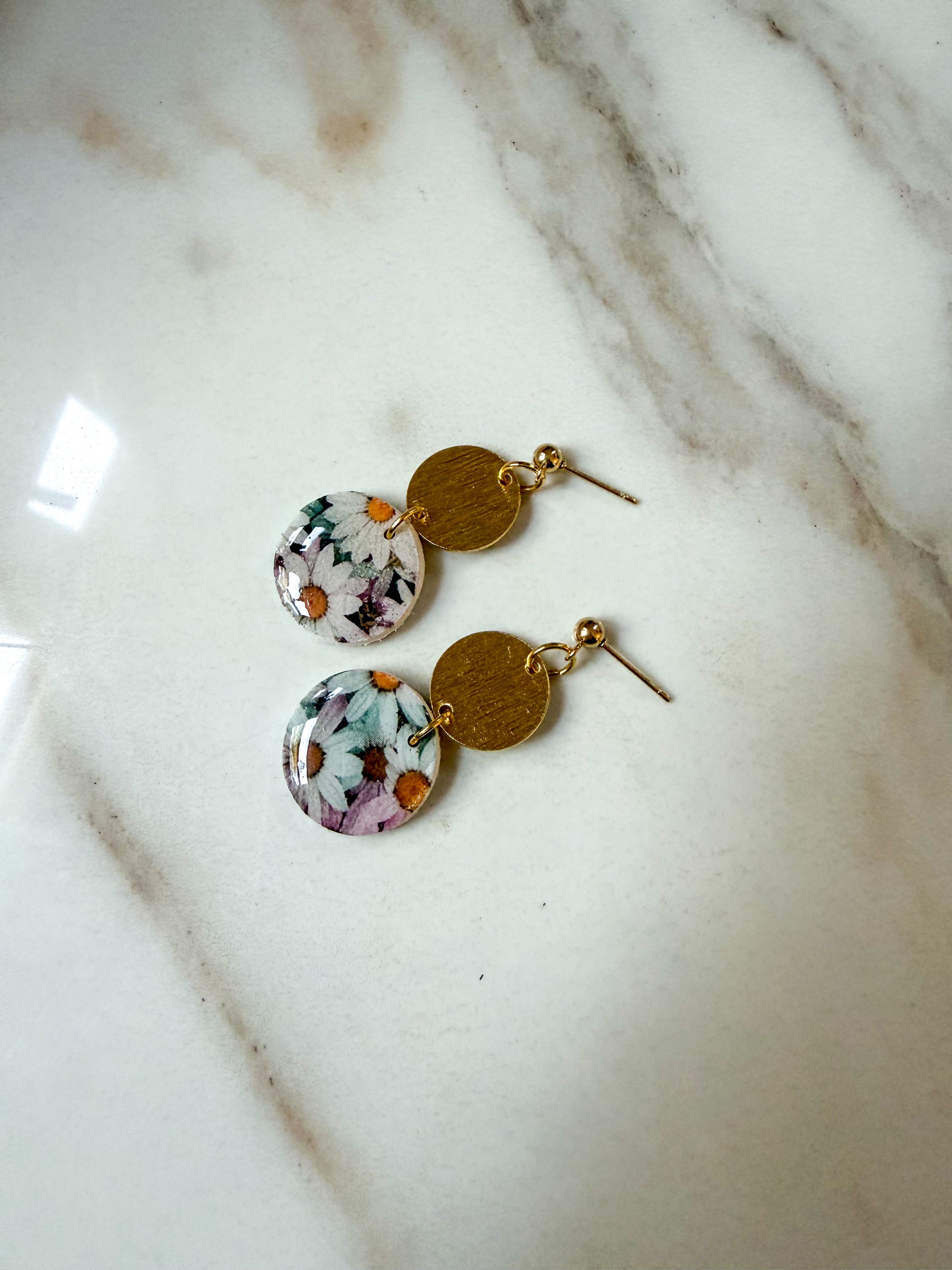 A pair of dangling earrings with round floral resin charms and gold-tone disc and post elements, displayed on a white marble surface.