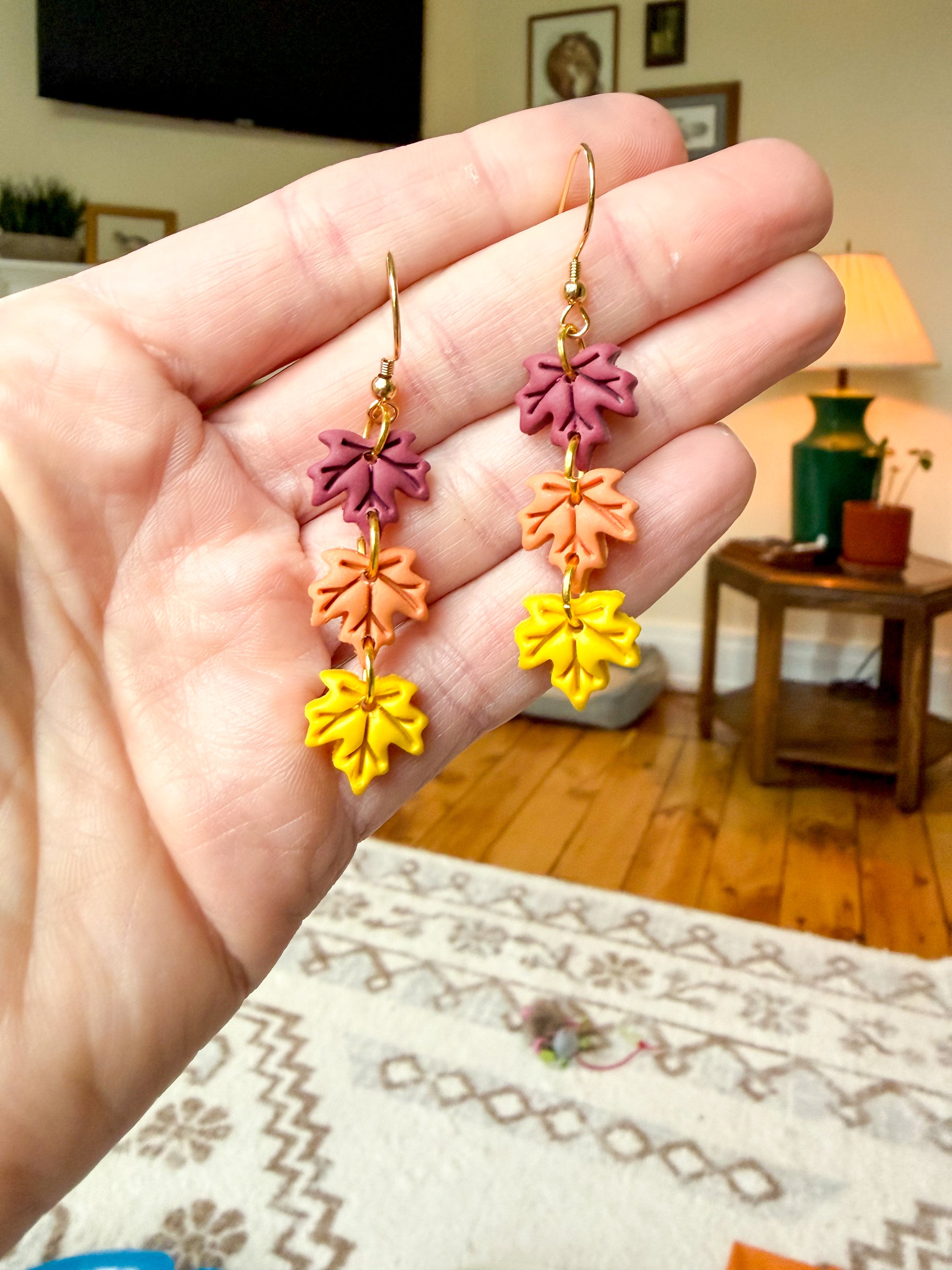 A person's hand holding a pair of gold-tone dangle earrings with three colorful leaf charms (purple, orange, and yellow) in an indoor home setting.