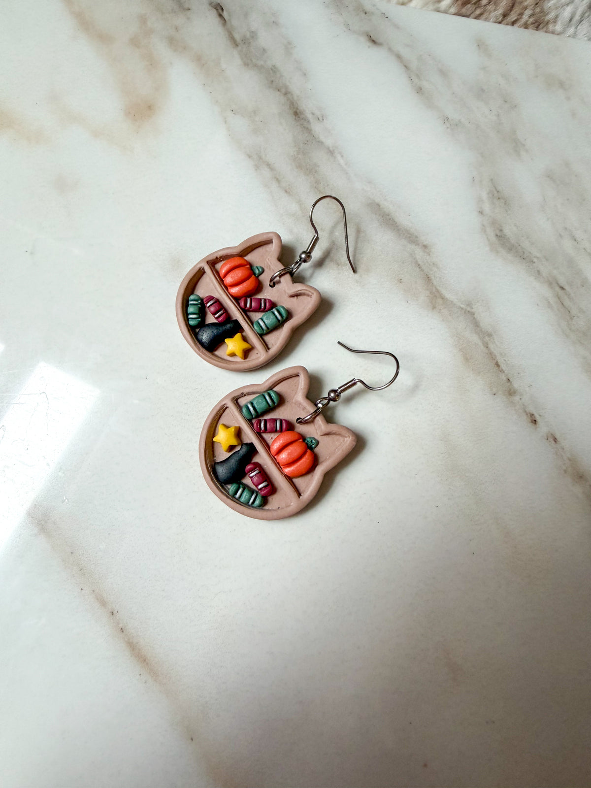 A pair of handmade cat-shaped dangle earrings featuring colorful book details and small orange pumpkin accents, resting on a white marble surface.