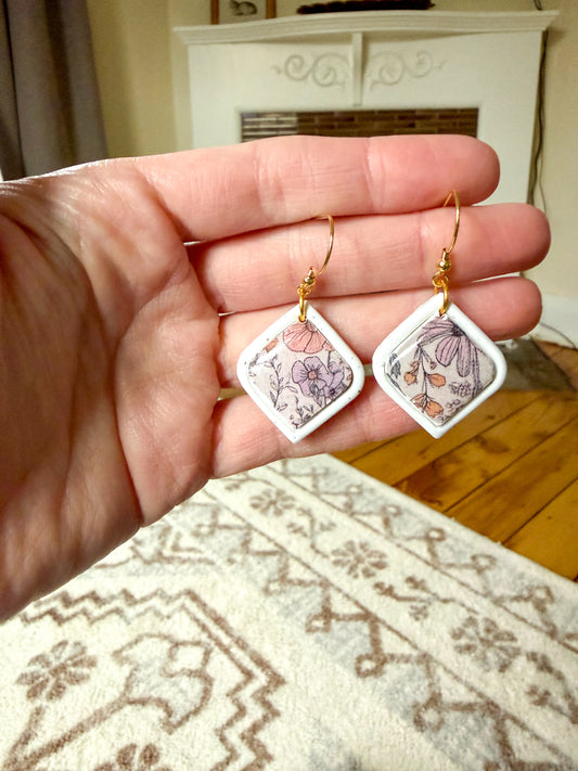 Floral earrings with a white frame held in a hand against a room background. 