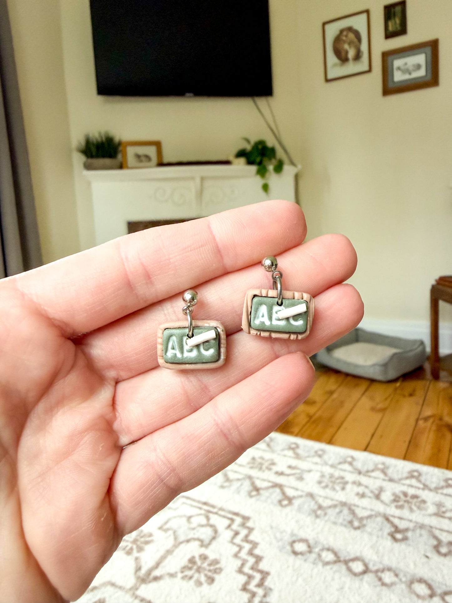 A hand holding a pair of earrings. Made to resemble little chalkboards with a piece of chalk. It reads ABC. 