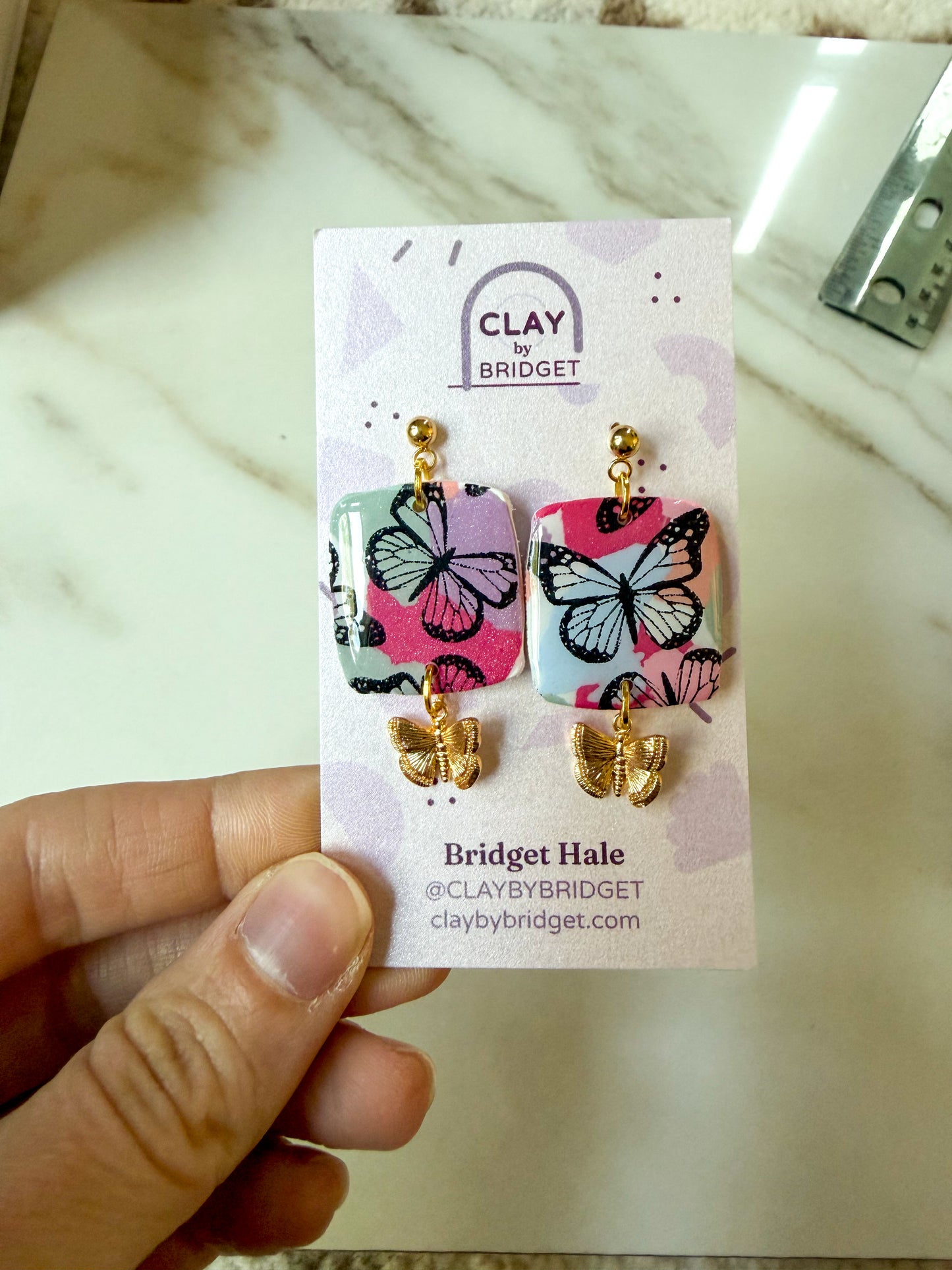Colorful butterfly dangles held in a hand. The earrings have a golden butterfly hanging from the bottom. 