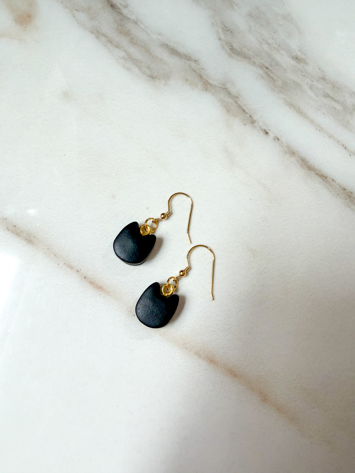A pair of black cat head shaped dangle earrings with gold fishhook wires and small gold bead accents, displayed on a white marble surface.