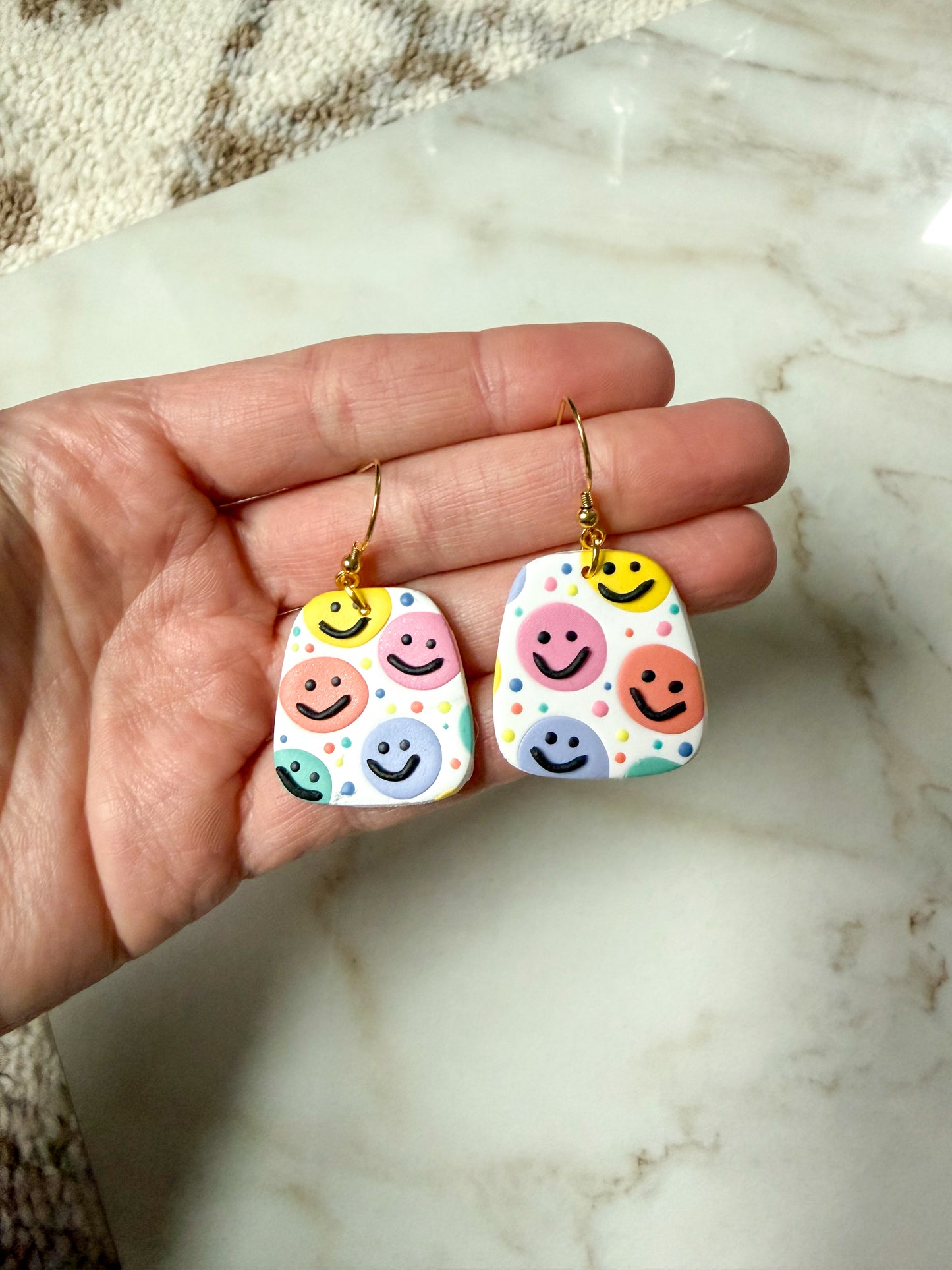 Colorful smiley face earrings held in a hand on a marble surface