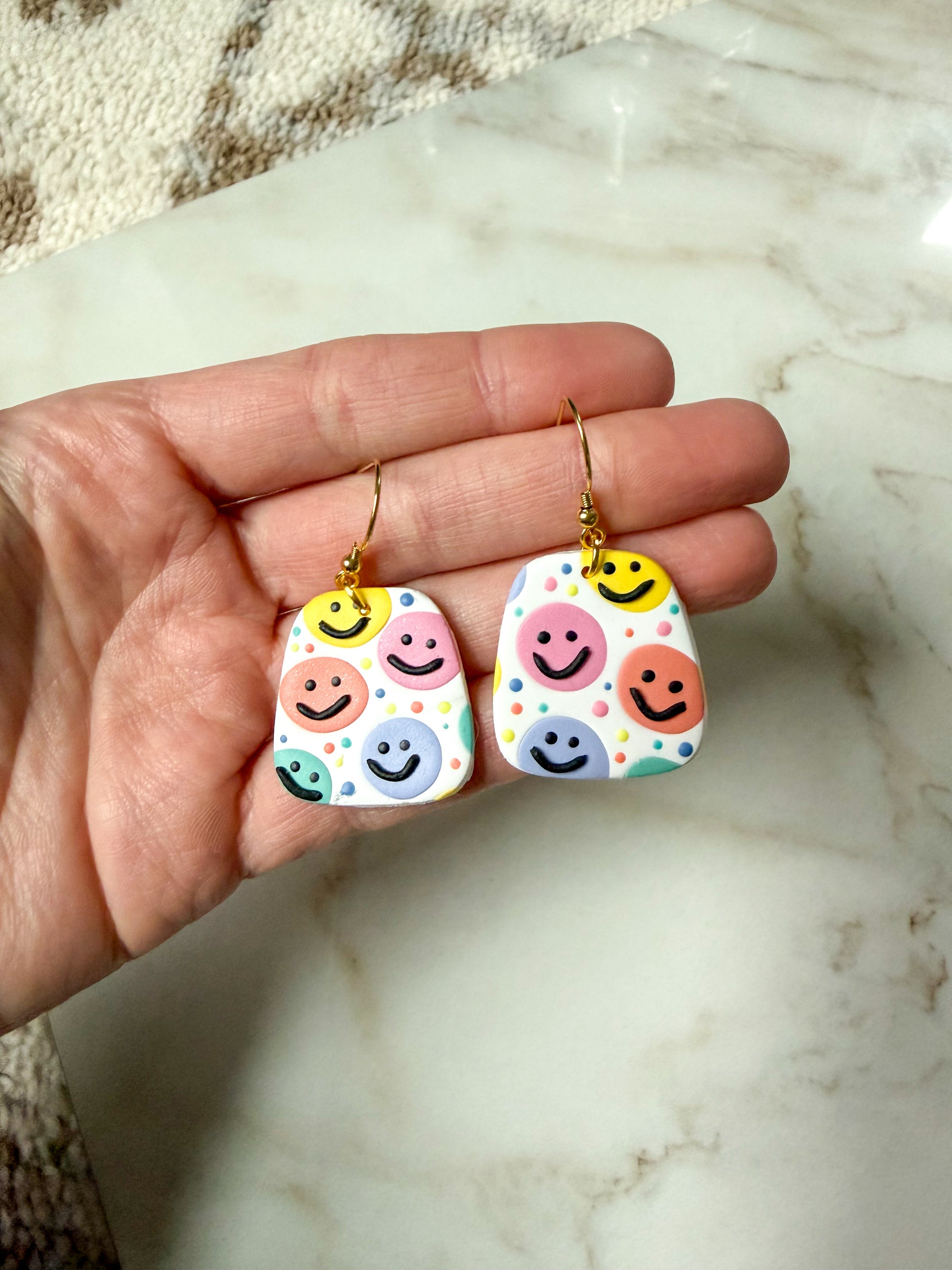 Colorful smiley face earrings held in a hand on a marble surface