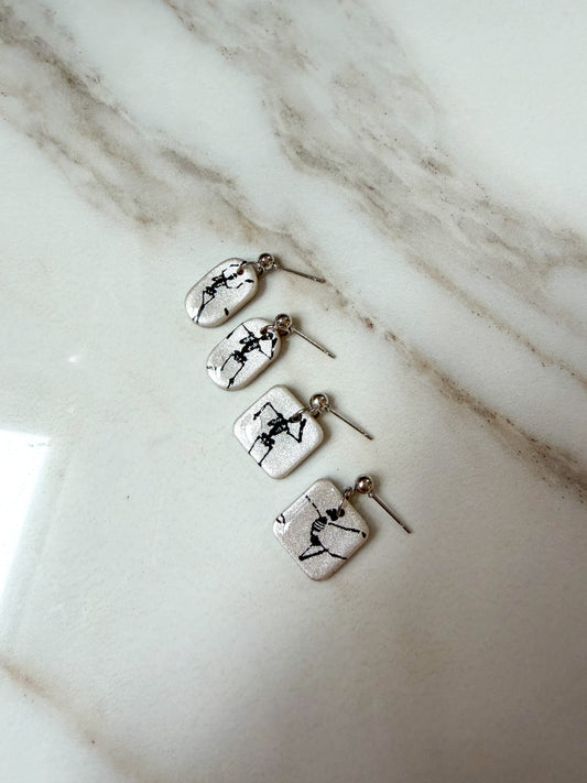 Four silver-toned earrings with small rectangular white pendants printed with black skeleton illustrations, arranged diagonally on a white marble surface.