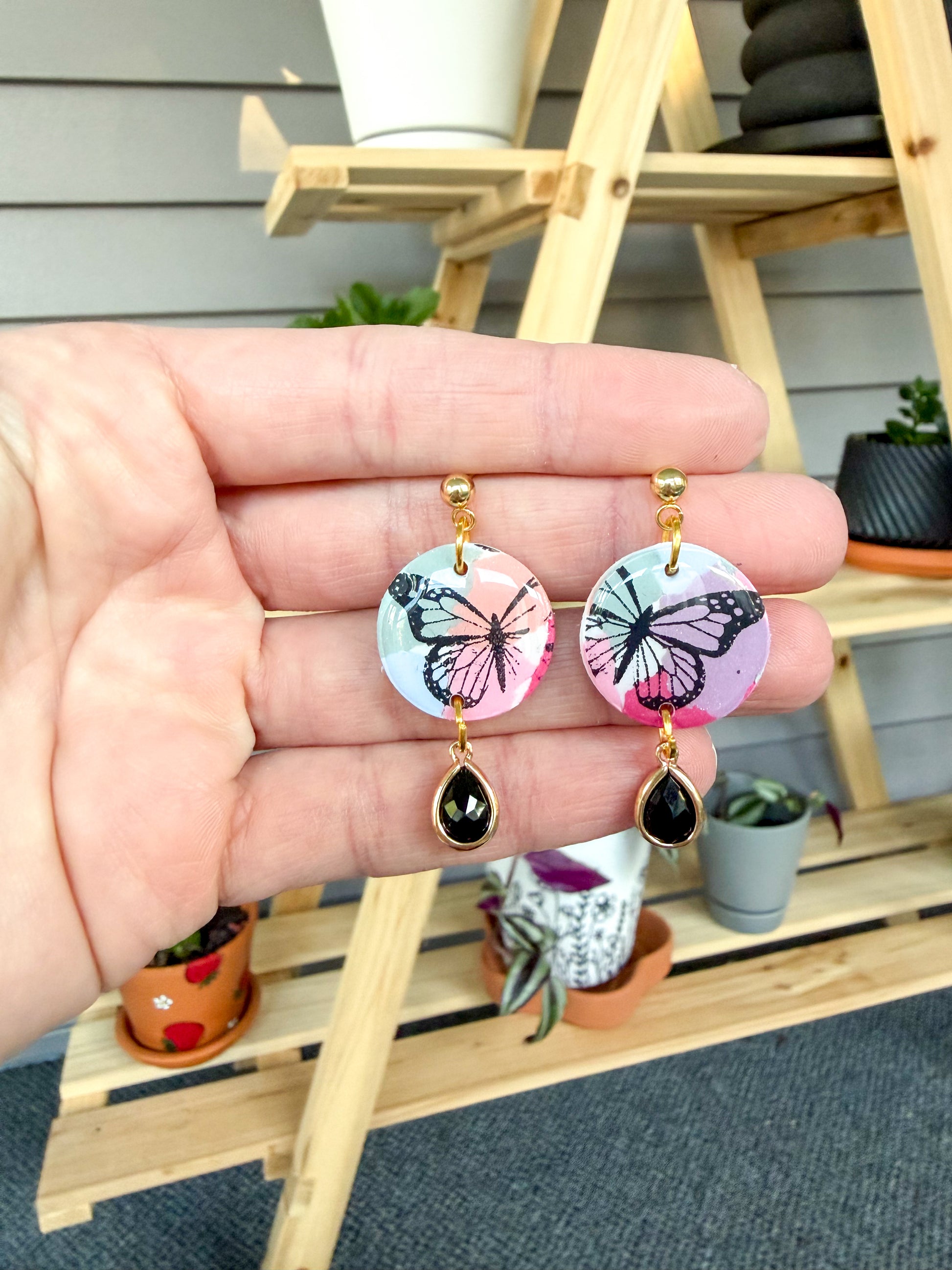Hand holding colorful earrings with a black butterfly stencil design, and a black charm hanging down. Held against a background with plants. 